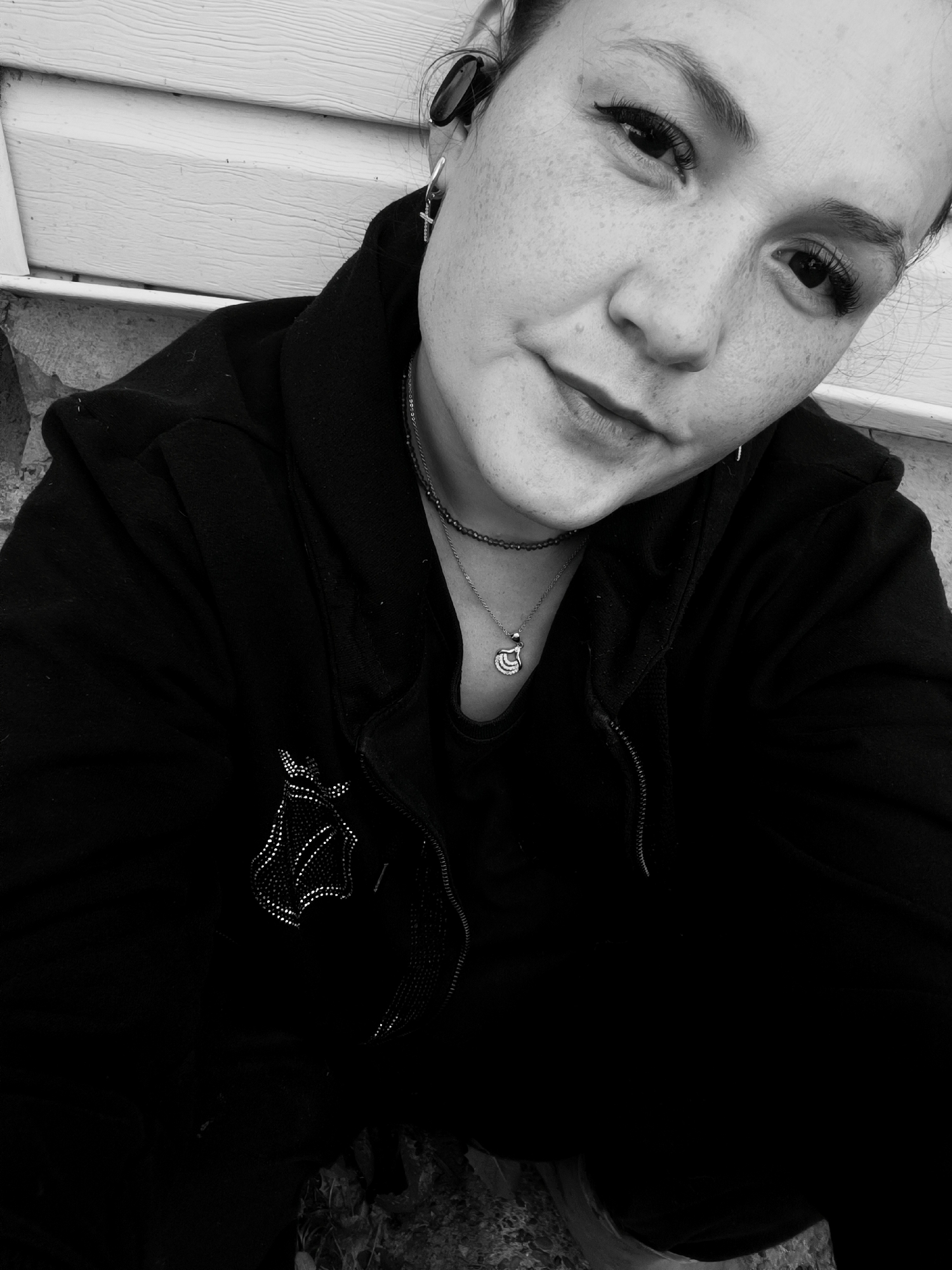 A black and white, close-up, eye-level selfie shows a young woman outdoors, with her face tilted towards the camera. She is wearing a black hooded jacket zipped partially, over a black t-shirt. Two necklaces are visible: one a thin, dark bead necklace and the other a silver chain with a shell-shaped pendant. She has noticeable freckles across her cheeks and nose, and is wearing dark eyeliner. A cross earring is visible on her left earlobe, and she has a black wireless earbud in her right ear. The background is out of focus, appearing to be a white siding wall of a building and some textured rocks or pavement. There is no discernible evidence in the image to suggest the location is in Karsha, Russia.