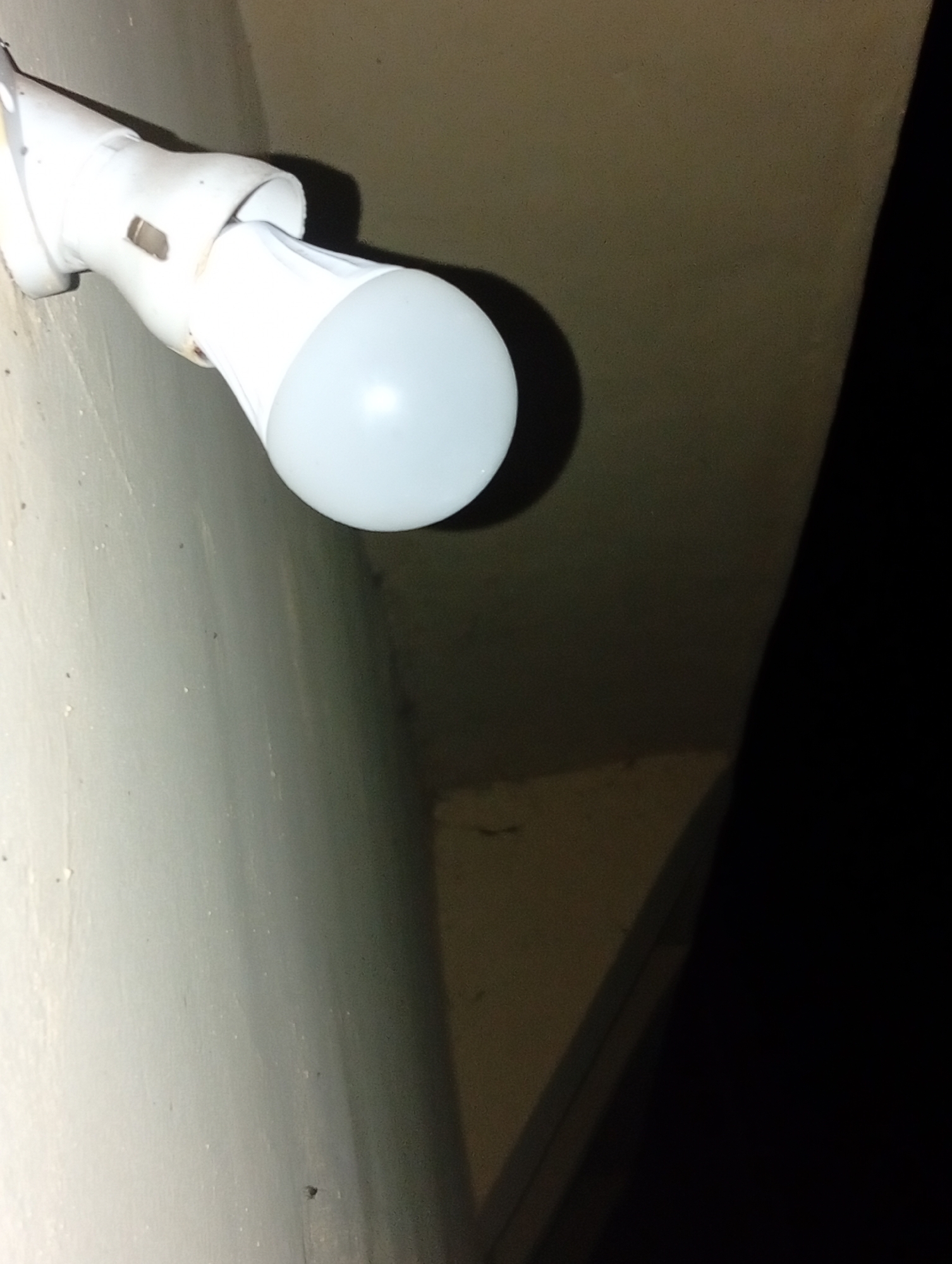 The image depicts a close-up, low-angle shot of a white LED light bulb screwed into a white light fixture. The light fixture is attached to a textured, light-colored wall, possibly concrete or plaster. The bulb itself is oval-shaped and emits a soft, diffused light. The background consists of a darker, textured surface, with a distinct shadow cast by the light fixture, suggesting an indoor environment with a light source positioned to create this shadow. The scene appears to be a dimly lit area. There is no indication of people, actions, or interactions. The context provided about Ukukwa, Nigeria, cannot be visually confirmed from the image itself.