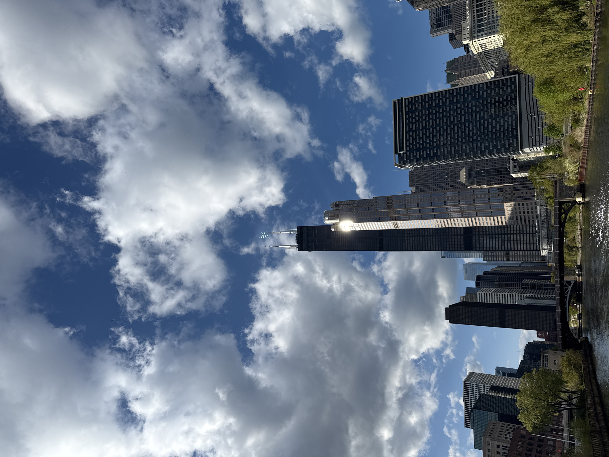 The image depicts a view in Chicago, United States, likely taken along the Chicago River. The foreground and right side of the image feature the river, its surface reflecting the sky and buildings. Along the riverbank, there are trees with green foliage and some grassy areas. The urban landscape consists of numerous skyscrapers of varying heights and architectural styles. Some buildings are modern, with glass facades, while others are older with more traditional designs. One prominent building is the Willis Tower (formerly Sears Tower), which is recognizable by its dark color and distinct stepped design. The sky is partially covered with white, puffy clouds, creating a mix of sun and shade. The overall impression is a bustling urban environment along a riverfront.