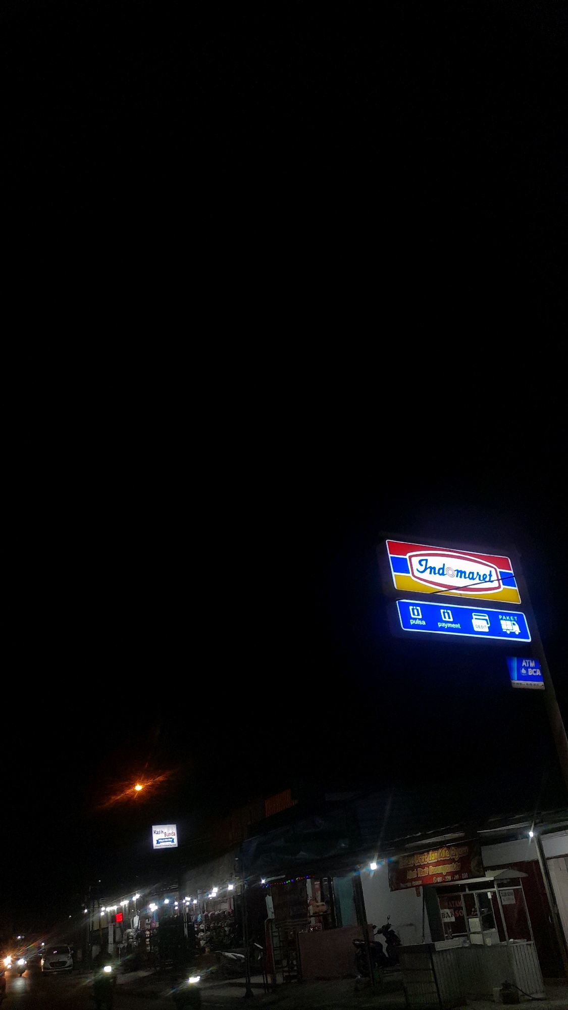 At night, the camera captures a street scene in Kalijeruk, Indonesia. A prominent Indomaret sign, illuminated with neon blue and red lights, is mounted on a pole. Below it, smaller blue signs indicate "pulsa," "payment," "debit," and "paket." A blue sign with "ATM" and "BCA" is also visible. Further down the street, there are several shops and market stalls, some of which are lit up. Vehicles, including a car and motorcycles, are present on the road. One motorcycle has a light on. The overall environment appears to be a commercial area with low-rise buildings and streetlights.