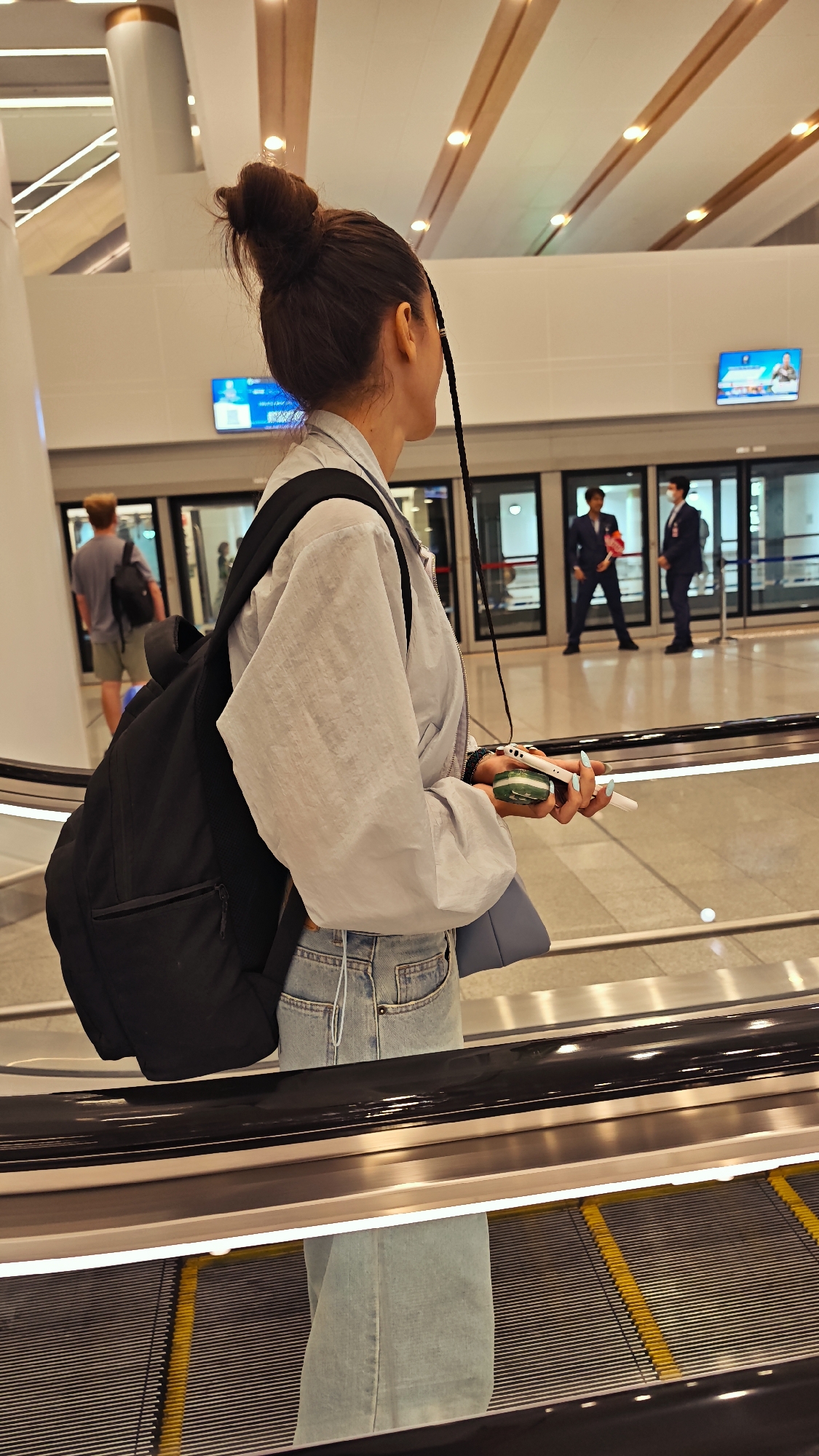 A young woman with dark hair pulled up in a messy bun and a single braid is standing on an escalator in what appears to be an airport terminal in Nong Prue Subdistrict, Thailand. She is wearing a light blue windbreaker jacket, light blue denim jeans, and a black backpack. In her right hand, she holds a white mobile phone and a small green object. Behind her, a man with short hair wearing a grey t-shirt and shorts with a backpack is walking away from the camera. Further in the background, two men in dark suits stand near automatic doors. The environment is modern and brightly lit with ceiling lights and digital displays.