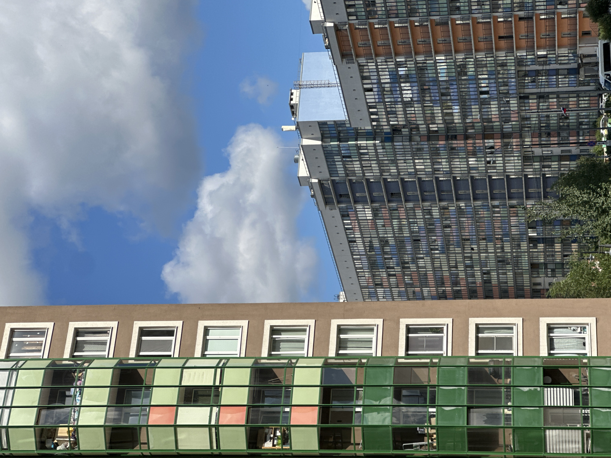 In the city of Murino, Russia, a photograph captures a section of a modern residential complex under a partly cloudy sky. On the left, a tall, rounded building with a green facade and numerous windows, some with balconies, stands prominently. To its right, a smaller, brown building with white-trimmed windows is visible. Further in the background, a much larger, multi-story building stretches across the frame, featuring a facade of numerous windows and balconies, with sections of blue, grey, and orange. The scene is dominated by the architecture of the buildings, set against a bright blue sky with scattered white clouds. No people or specific actions are discernible in the image.