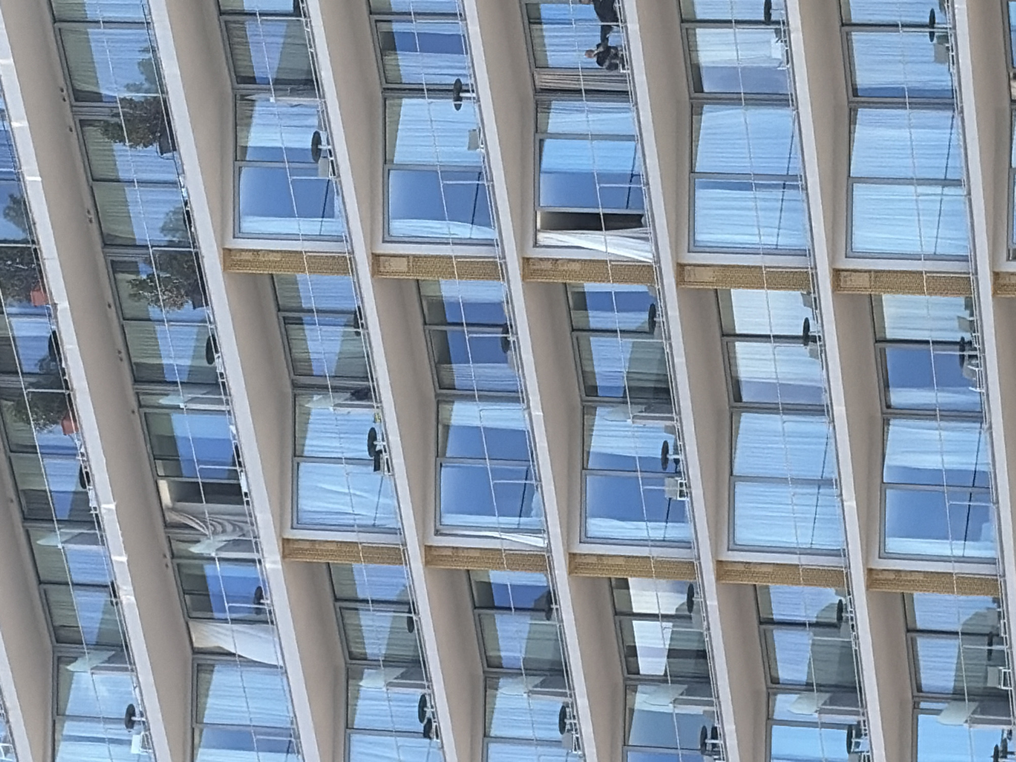 The image depicts a section of a building exterior in Bečići, Montenegro, notable for its repeated pattern of windows. The windows are open slightly, allowing glimpses of either interior spaces or the sky. Some windows are fitted with light-colored blinds. The building's architecture features light-colored frames that accentuate the pattern, creating a rhythmic visual. Reflections are visible in the window panes. From one of the windows, you can see a man walking, indicating the location is occupied.
