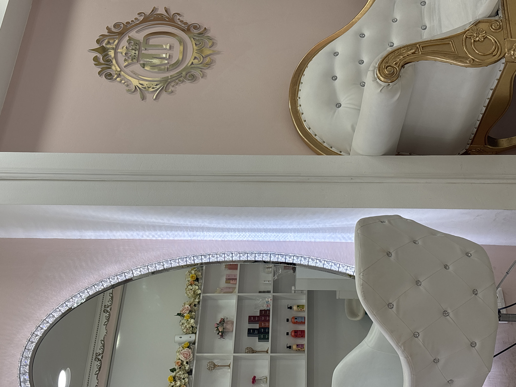 This media file captures an opulent indoor setting, likely a high-end salon, boutique, or beauty establishment located in Sofia, Bulgaria. The image is composed with a horizontal white architectural beam or ledge dividing the frame.
The upper portion of the image displays a pale pink wall adorned with a prominent, ornate golden emblem. This decorative piece features a stylized crown positioned above a monogram, which appears to intricately intertwine the letters 'G' and 'E' or 'P'. To the right, a segment of a luxurious white armchair is visible, showcasing a golden carved frame and deeply tufted upholstery studded with sparkling, jewel-like buttons.
The lower part of the image is dominated by a large, possibly oval or arched mirror, distinguished by its glittering frame encrusted with numerous clear, rectangular crystal or jewel-like elements. The mirror reflects a section of the room, revealing a white shelving unit filled with various beauty products, including bottles and boxed items. Decorative floral garlands, consisting of yellow and pink roses, cascade down the side of the shelving unit. Also reflected is the back and side of another white, tufted chair, matching the opulent style of the one seen in the upper half of the image.
The overall aesthetic conveys an atmosphere of elegance, luxury, and comfort. No people are present in the scene, and no specific activity is taking place. The lighting suggests an indoor environment, with no discernible cues for the time of day or external weather conditions.