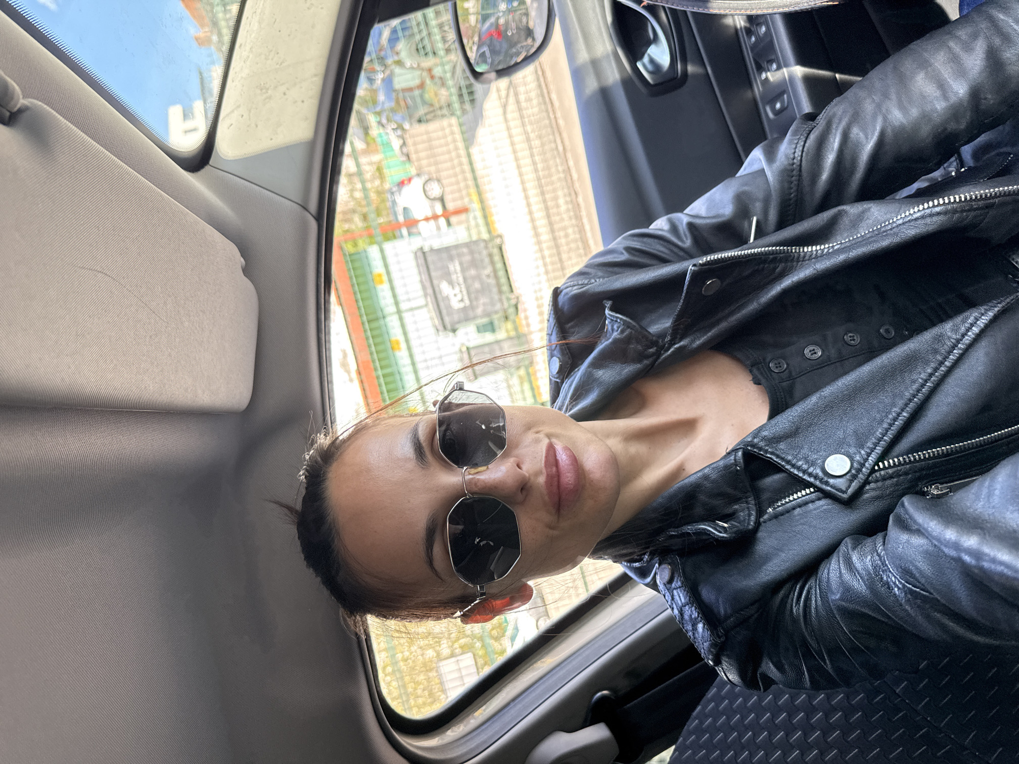The image captures a woman posing for a photo inside a car during the daytime in Pleven, Bulgaria. She is seated, likely in the passenger seat, and looks directly at the camera with a relaxed, confident expression and a slight smile. She is wearing stylish hexagonal/octagonal sunglasses and a black leather jacket featuring visible zippers and metal snaps. Her dark hair is pulled back.
The car's interior is visible, showcasing a grey sun visor, grey roof lining, and a patterned black car seat. Through the car window, a bright outdoor scene is discernible under a clear blue sky, indicating sunny weather. The background suggests an urban or suburban environment with buildings and structures. Among these, a white structure resembling an 'H' shape is faintly visible, and behind it, some green, scaffold-like elements can be seen. There is also a blurry black rectangular sign with indistinct white text on it. The bright natural light illuminates the scene, highlighting the details of the woman's attire and the car's interior.