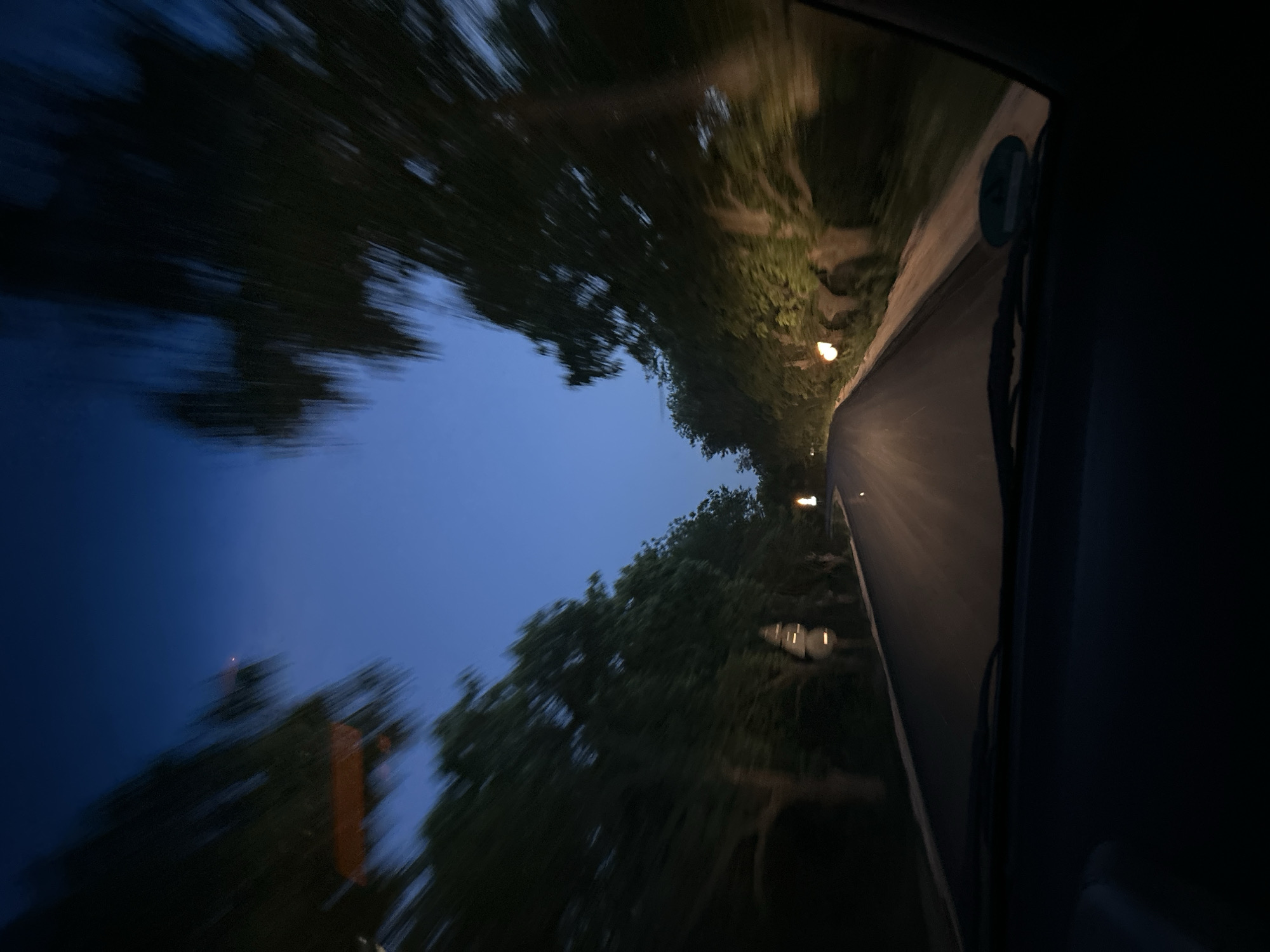 The image appears to be taken from inside a moving vehicle at dusk or night. The view is primarily of a road flanked by trees or vegetation, likely in a rural or suburban area. Streetlights are visible along the road. There is considerable motion blur, suggesting that the vehicle is traveling at speed. The location is unknown. There are no people visible in the image.