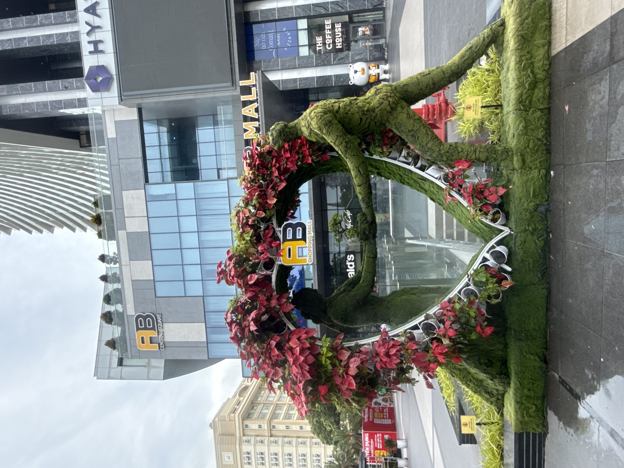 The image depicts a street-level view in front of a modern commercial building complex in Nha Trang Ward, Vietnam. The primary subject is a large, heart-shaped floral and topiary sculpture. The sculpture is composed of green, moss-like material forming human-like figures on either side, holding hands, and enclosing a reflective surface. The heart shape is further adorned with vibrant red poinsettia-like flowers and greenery.
Behind the sculpture, the building facade features glass windows and signage. Prominent signs include "AB CENTRAL SQUARE" and "MALL" on one section, and "HYATT" on another. The complex appears to be a shopping mall and hotel. In the background, a taller, modern building with a striped facade is visible. To the left of the main building, a lighter-colored, multi-story building with numerous windows stands.
On the ground, the pavement is wet, suggesting recent rain. There are some yellow signs placed in front of the sculpture. A red fire hydrant is visible near the base of the sculpture. A small statue of a white bear with a yellow vest is positioned to the right of the main sculpture, near the entrance to "THE COFFEE HOUSE". The sky is overcast. A few people are partially visible in the distance walking in front of the buildings.