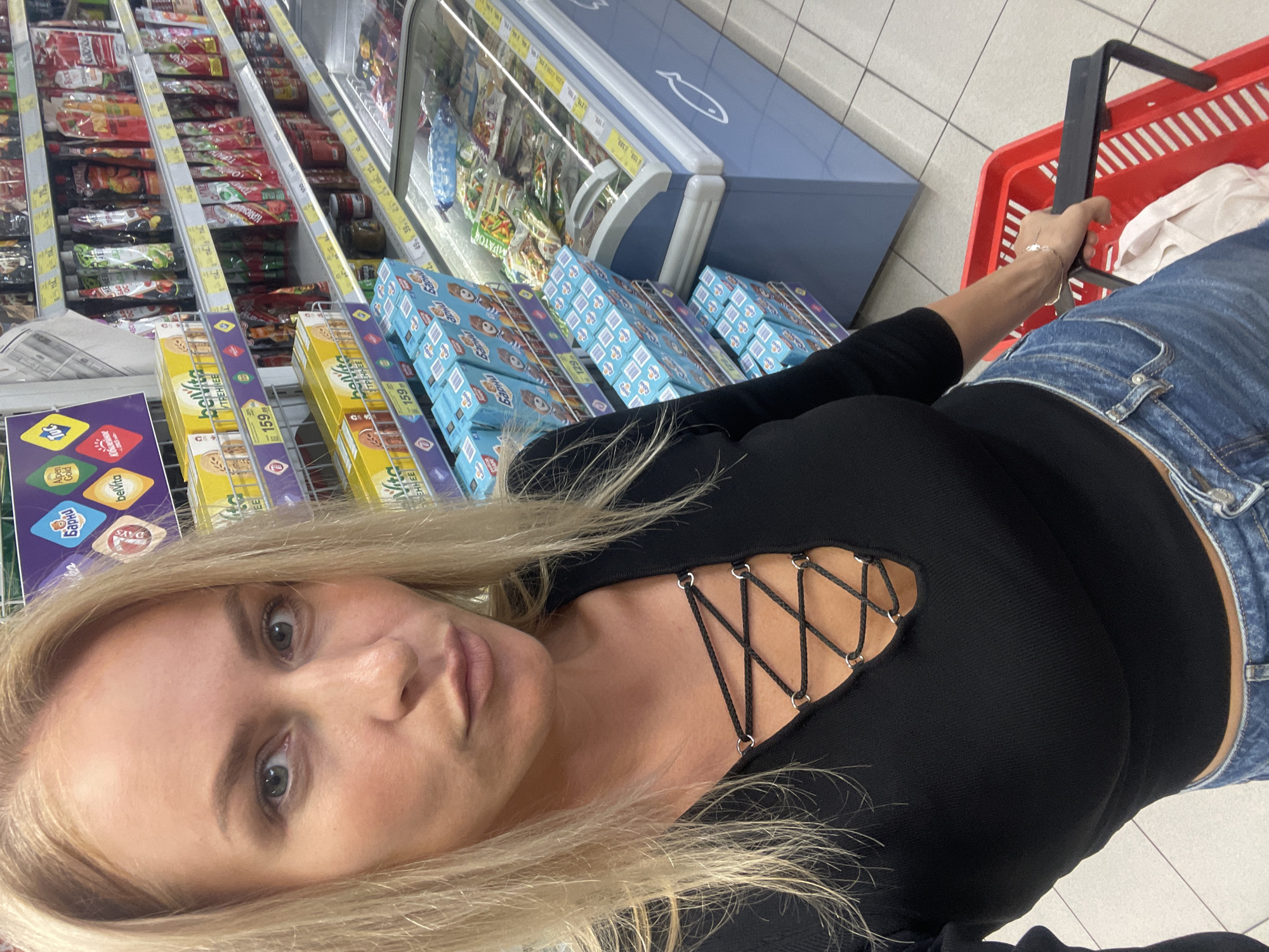 The image shows a blonde woman taking a selfie in what appears to be a grocery store. She is wearing a black top with lace-up detailing at the neckline and blue jeans. She is holding a red shopping basket. Shelves stocked with various food items and refrigerated display cases can be seen in the background. The labels on the products are in Russian, and prices are visible, indicating the location is likely in Russia. Based on the available text and context, it is unlikely to be possible to determine if it is specifically in Baranovka, Russia.