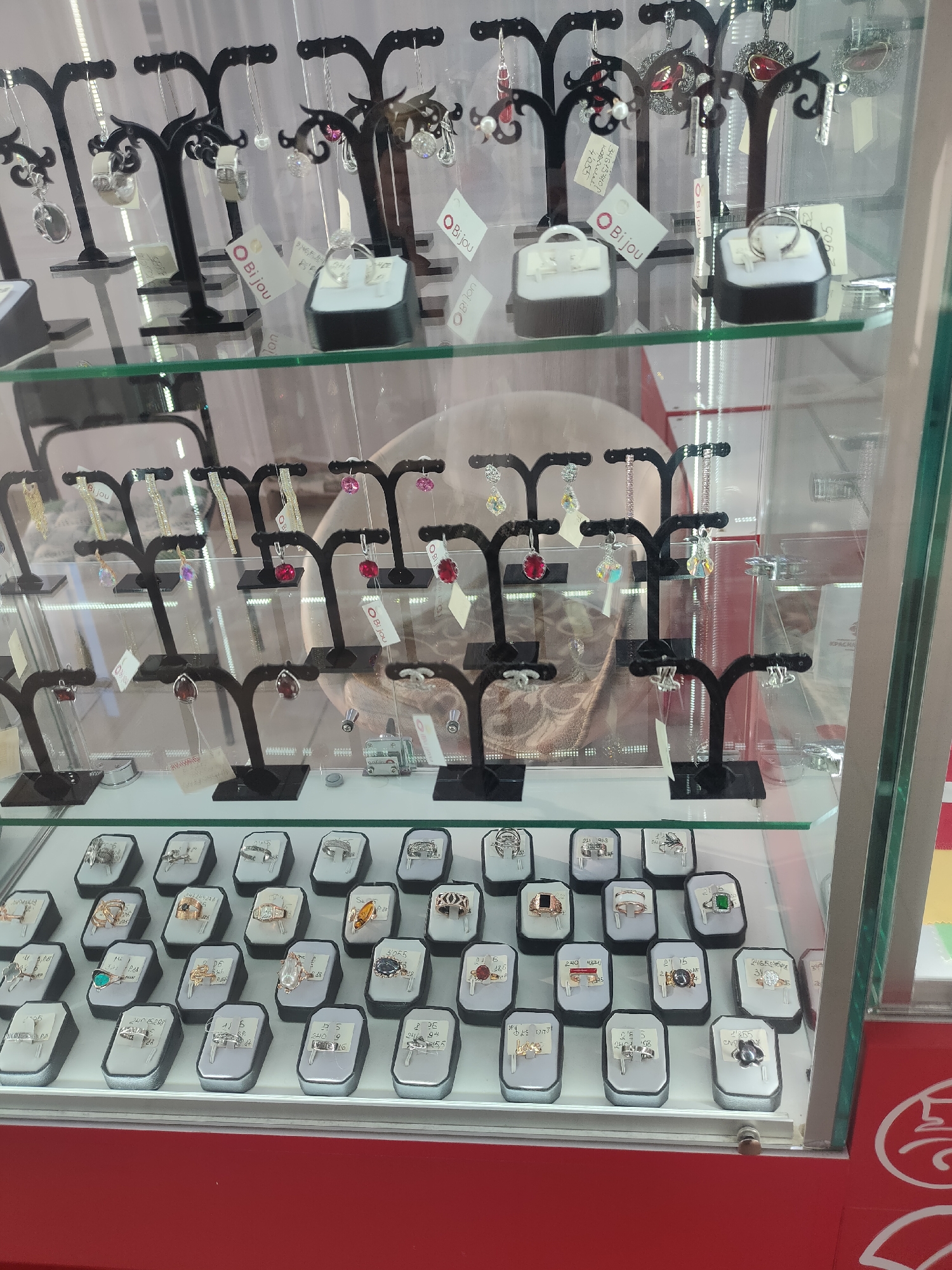 The image shows a display case in a jewelry store in Oktyabrsky, Russia. The display case is filled with various pieces of jewelry, including necklaces, earrings, and rings. The jewelry is arranged on black stands and white trays. The display case is made of glass and is lit from within, illuminating the jewelry. The background of the image is blurred, but it appears to be a store interior with red walls and a white ceiling. There are no people visible in the image.