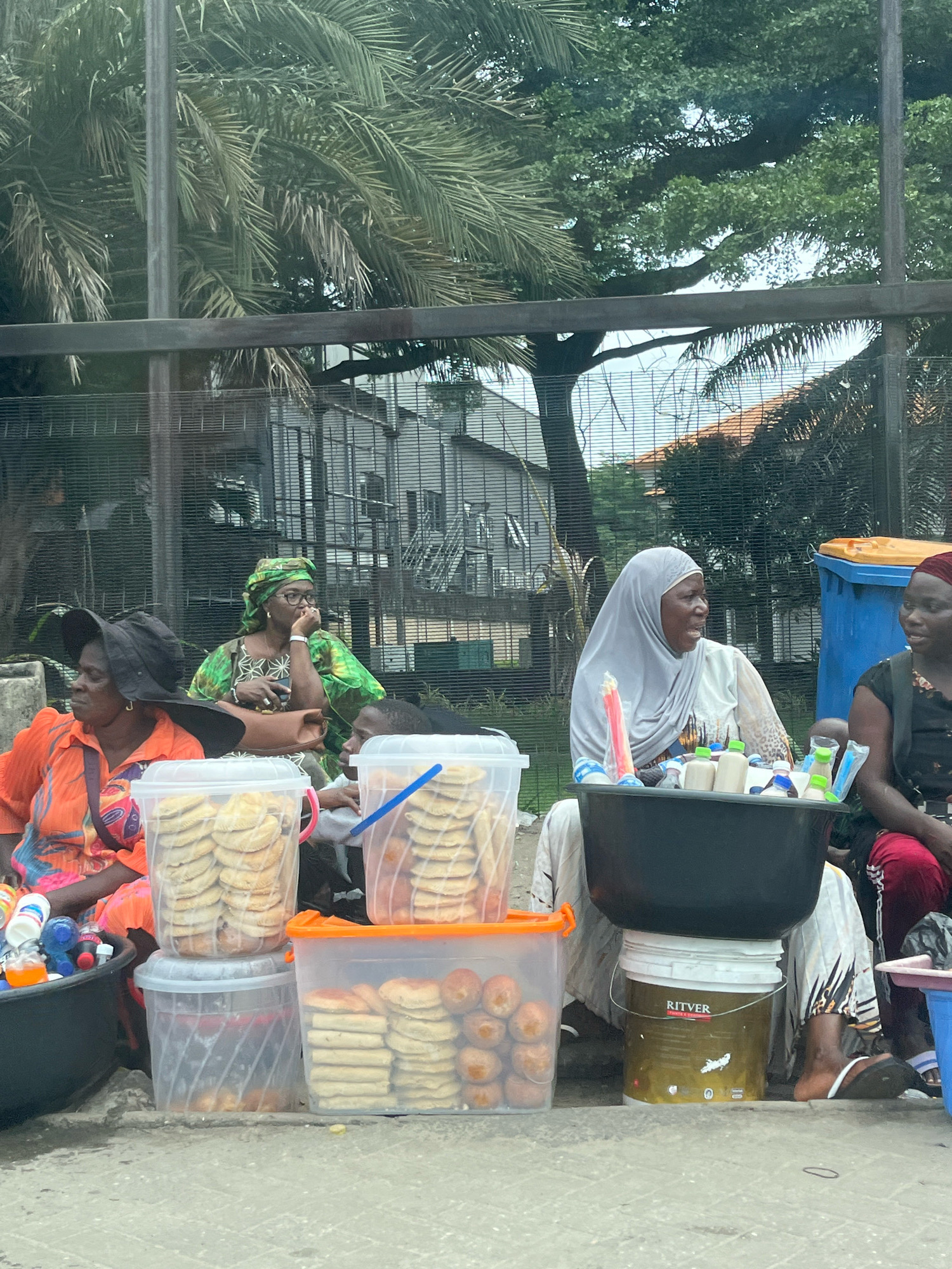 In Lekki Phase I, Nigeria, several women are seen selling food items from their roadside stalls. The scene depicts a vibrant street market with an array of goods displayed in plastic containers and buckets. In the foreground, there are large clear plastic buckets filled with what appear to be baked goods, likely cookies or pastries. Next to these, another woman is selling beverages and other small items from a large black basin balanced on a paint bucket. The women are dressed in traditional African attire, with headscarves and colorful fabrics being prominent. They are engaged in conversations with each other and possibly customers, though no customers are clearly visible interacting with the food stalls. Behind them, a metal fence separates the market area from a building and lush green trees, including palm trees, which suggest a tropical or subtropical environment common in coastal Nigeria. The ground is paved, indicating an urban setting. The overall atmosphere appears to be one of daily commerce and community interaction.