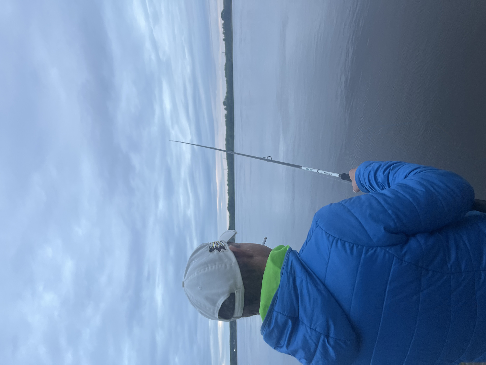 An individual, viewed from behind, is depicted on the bank of a large body of water, likely the Votkinsk Reservoir, in Chaikovsky, Russia. The person is wearing a light-colored baseball cap with a dark emblem on the right side and a bright blue hooded puffer jacket, revealing a neon green collar or hood liner underneath. A lit cigarette is visible protruding from the right side of the individual's head. The right arm holds a white and grey fishing rod, labeled "MARK" and "NAUTILUS," which extends towards the calm, dark water. In the midground, a distant shoreline lined with green trees is visible across the water, with faint industrial structures discernible above the tree line. The sky is overcast with a mix of grey and light blue clouds.