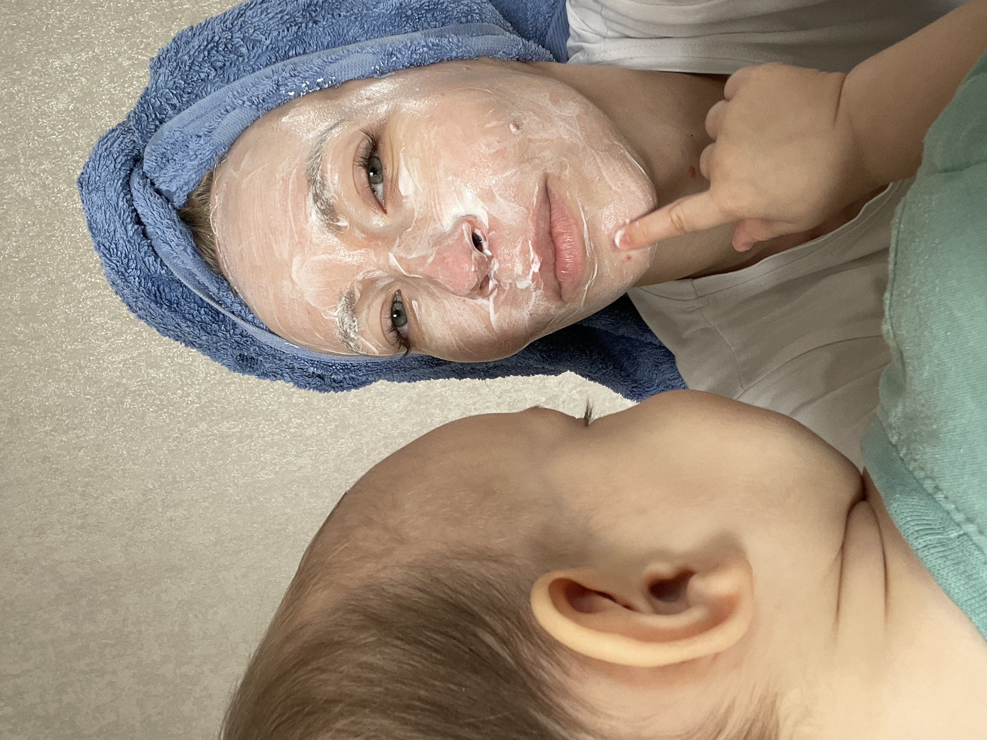 A medium close-up shot depicts a woman and a baby. The woman is lying down with a blue towel wrapped around her head. Her face is covered with a white cosmetic mask. A baby, lying next to her, is pointing at the woman's cheek with its right index finger. The baby is wearing a green shirt. The background is a beige wall. There is no indication of the location being Perm, Russia.