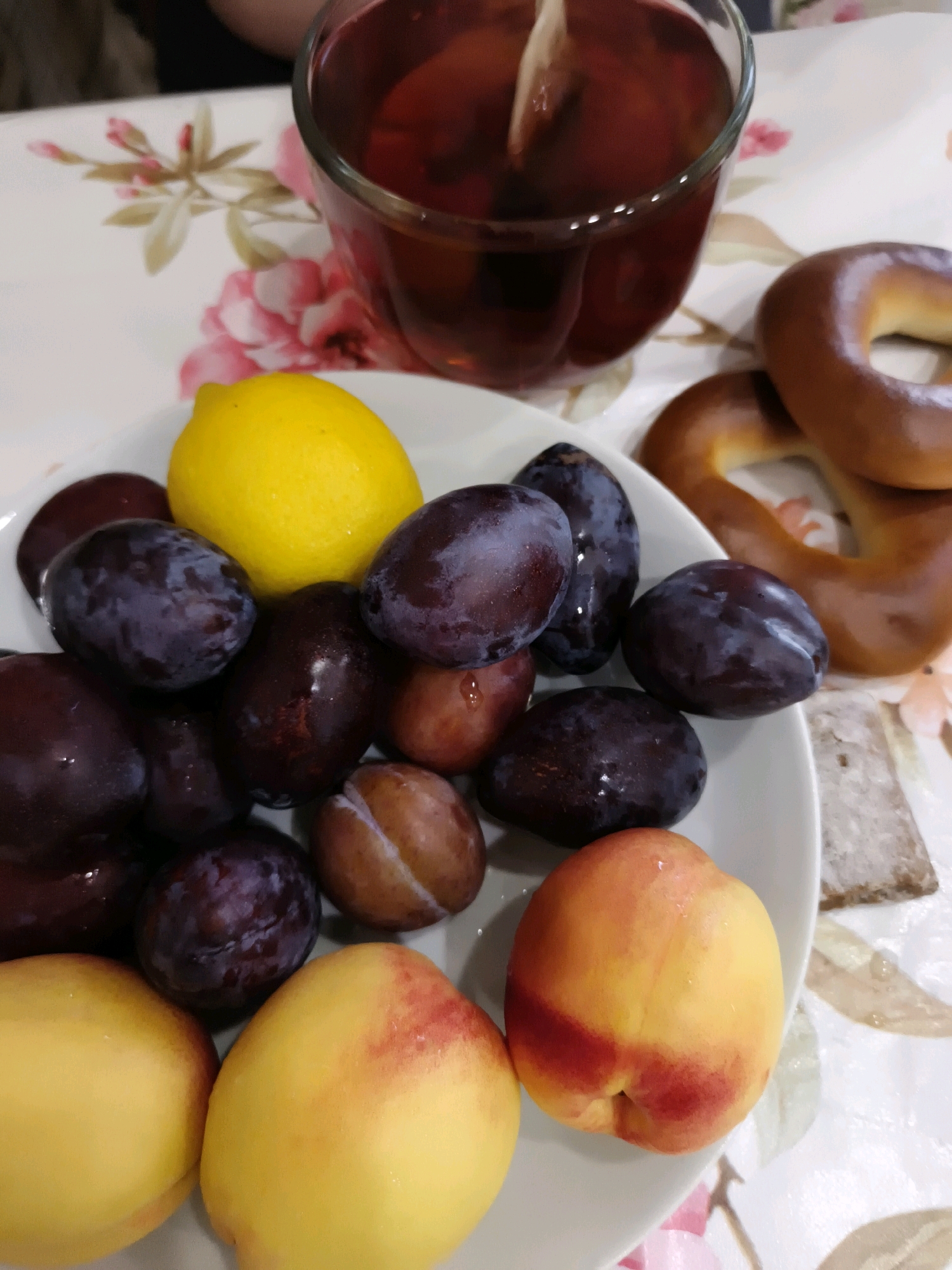 This is a close-up, eye-level, indoor shot of a platter of fruit and other food items, possibly on a table covered with a floral-patterned tablecloth. The subjects are arranged in the foreground and midground.
In the foreground, a white, round plate holds a collection of fruits. Two ripe peaches, one on the left and one on the right, are prominent. They are yellow with red blushes. Scattered among them are numerous dark purple plums, some appearing plump and glossy. A bright yellow lemon sits atop the plums in the upper left of the plate.
In the midground, behind the plate of fruit, a glass of dark red liquid, likely tea, with a tea bag inside is visible. To the right of the tea, two golden-brown pretzels are positioned on the tablecloth. A small, rectangular, light-brown object, possibly a cracker or piece of bread, is also present on the tablecloth.
The background is largely out of focus but suggests an indoor setting. A hint of a person's shoulder and arm, wearing dark clothing, is visible in the upper left corner. The floral pattern on the tablecloth features pink and green elements.
The image does not contain any specific visual cues that would indicate its location in Yoshkar-Ola, Russia. The scene depicts a casual food setting.