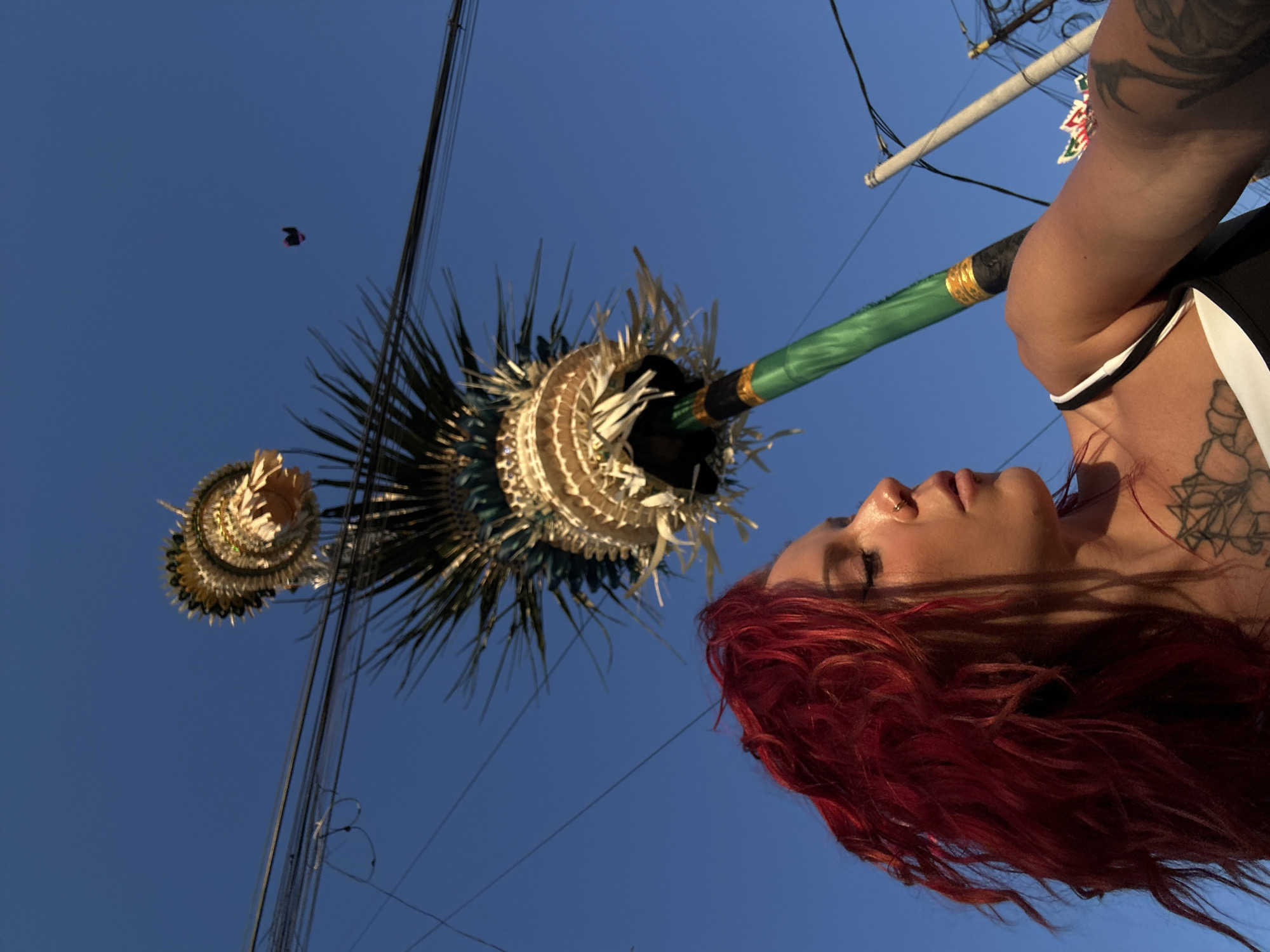 In North Kuta, Indonesia, a woman with vibrant red hair is captured in a low-angle shot, looking upwards and to the right with her eyes closed. She is wearing a black and white tank top, revealing tattoos on her chest and arm. She has a nose ring. She is holding a tall, decorative pole with elaborate woven decorations and feathery accents, possibly for a ceremony or festival. The pole is green with gold accents near the top. The background is a clear blue sky with power lines crisscrossing it. A small dark object, possibly a kite, is visible in the distant sky. The scene suggests an outdoor cultural event.