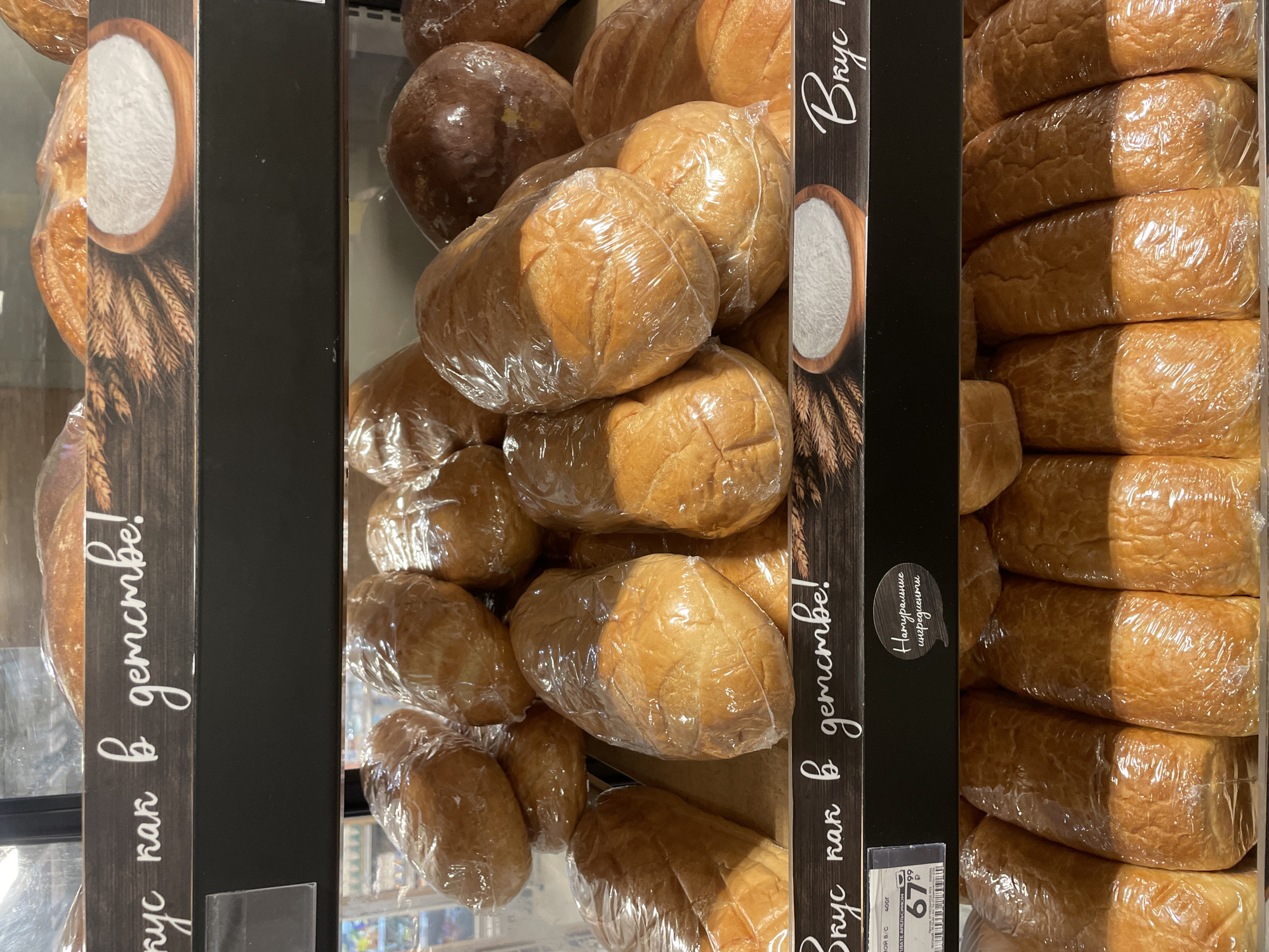 The image shows shelves stocked with bread in what appears to be a grocery store in Moscow, Russia. The bread on the left and right are wrapped in plastic and arranged in rows. The bread in the center are wrapped with plastic but are in piles. Each section is marked with a label that says “Вкус как в детстве!” which means "A taste like in childhood!" There is also a sticker that says "Натуральные ингредиенты" meaning "Natural ingredients." The price tag for the bread in the right corner is 67.89 Russian rubles.