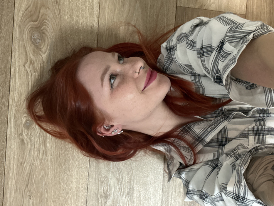 A young adult female, appearing to be in her 20s or 30s, is depicted lying supine on a light-colored wooden floor in an indoor setting in Rostov-on-Don, Russia. Her head is positioned towards the upper left quadrant of the frame, with her body extending diagonally downwards. She has long, wavy, bright red hair spread out around her head. Her face features light-colored eyes with dark eyeliner, visible freckles, and pink lipstick. She exhibits a slight smile, with her gaze directed upwards and to the right from the viewer's perspective.
The individual has multiple body piercings: a black circular barbell in her nasal septum, and several piercings on her visible left ear, including a crescent moon-shaped earring in the helix/conch area, a floral cluster earring below it, and a hoop in the lobe. She is dressed in a white and black plaid button-up shirt, with the sleeve on her visible right arm rolled up, revealing a dark tattoo on her forearm. The environment consists solely of the light brown wooden floor, characterized by visible grain patterns and seams between planks. There are no other individuals or prominent objects in the frame, and no direct interactions are depicted.