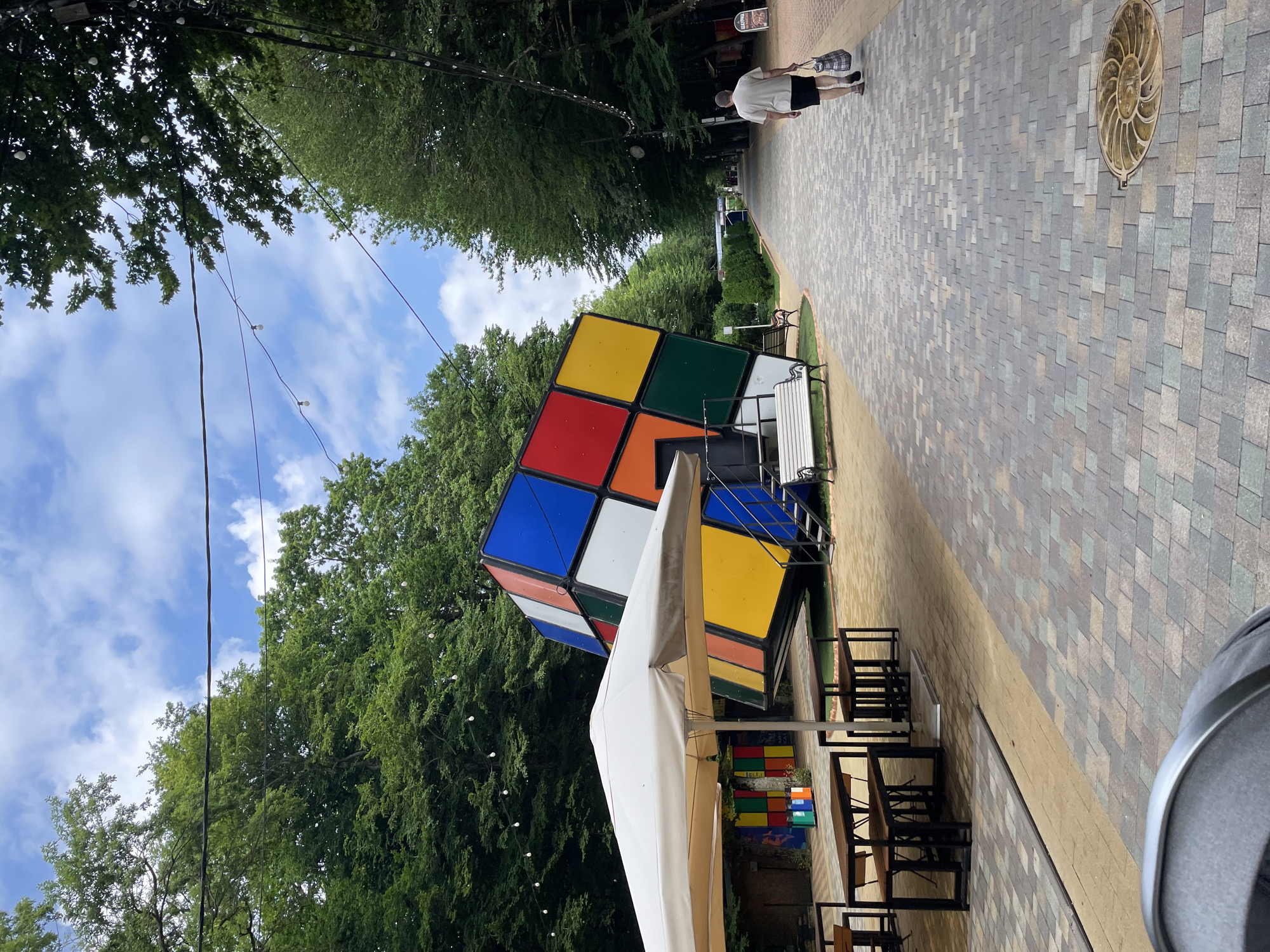 The image depicts an outdoor area featuring a large, tilted Rubik's Cube sculpture as its focal point. The sculpture is situated in what appears to be a park or recreational space, surrounded by lush green trees. A paved walkway runs alongside the sculpture, leading towards a distant person walking away from the camera, carrying a bag. Several outdoor tables and chairs are visible under a white umbrella, suggesting a seating area. String lights are hung between trees, creating an ambient atmosphere. In the background, more trees and some buildings can be seen. The sky is partly cloudy. The location is identified as Stavropol, Russia.