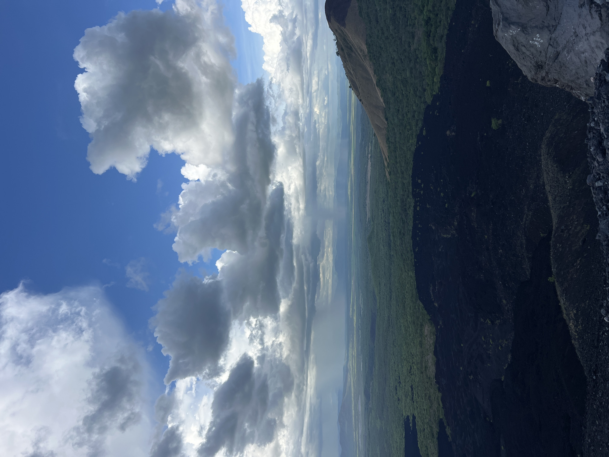 An eye-level, wide-angle, vertical shot depicts a vast, dramatic landscape dominated by dark volcanic rock in the foreground and a lush green expanse stretching to the horizon under a dynamic sky. The immediate foreground is a rugged, uneven terrain composed of dark, porous volcanic rock, with a few scattered grey rocks and a larger, rough-textured grey boulder on the right edge of the frame. Beyond this, a dense, dark green forest covers a gently sloping area, transitioning into a flatter, lighter green plain that extends to distant, hazy blue mountains. A patch of rain is visible as a grey, misty curtain in the middle ground, suggesting a weather event in progress. The sky above is a mix of deep blue and large, billowing grey and white clouds, with some areas of brighter white suggesting sunlight breaking through. The overall impression is one of raw, natural beauty and geological activity, possibly from an elevated vantage point overlooking a volcanic crater or lava field. The location is identified as Masaya Volcano National Park, Nicaragua.