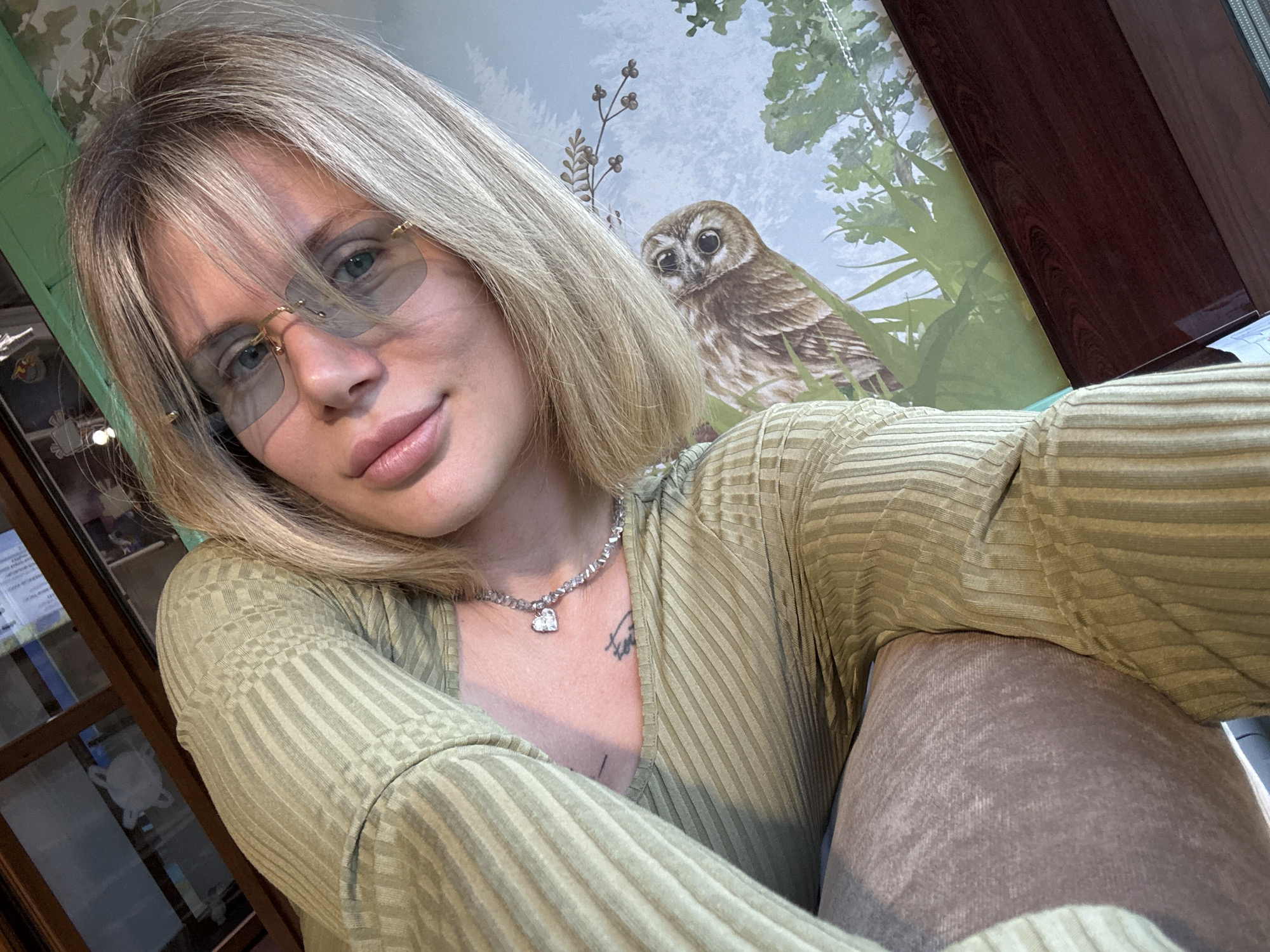 A selfie-style portrait captures a young woman looking directly at the camera with a soft, relaxed expression. She has blonde hair with bangs falling across her forehead and light-colored eyes. She wears rimless sunglasses with light grey-tinted lenses and delicate gold frames. Her lips are full, showing a slight smile.
She is dressed in a long-sleeved, light olive green top featuring a ribbed texture. A silver-colored necklace with a sparkling heart-shaped pendant adorns her neck, and below it, a tattoo with indistinct cursive text is visible on her chest. In the foreground, to the lower right, part of her leg is visible, covered in light brown, ribbed fabric, possibly indicating pants or a blanket.
The background is an indoor setting with decorative wallpaper. The most prominent feature of the wallpaper is a brown owl with large, dark eyes, perched on a branch and looking forward. Surrounding the owl are illustrations of green trees, foliage, and small berry-like plants, creating a natural scene. To the left, a dark-framed glass cabinet or shelving unit is partially visible, containing blurry objects. On the far right, a dark brown, possibly wooden, surface provides a contrast to the wallpaper. The lighting suggests daytime. No specific text is clearly legible in the image, although there might be blurry text on the tattoo and within the cabinet. The overall mood is calm and casual.