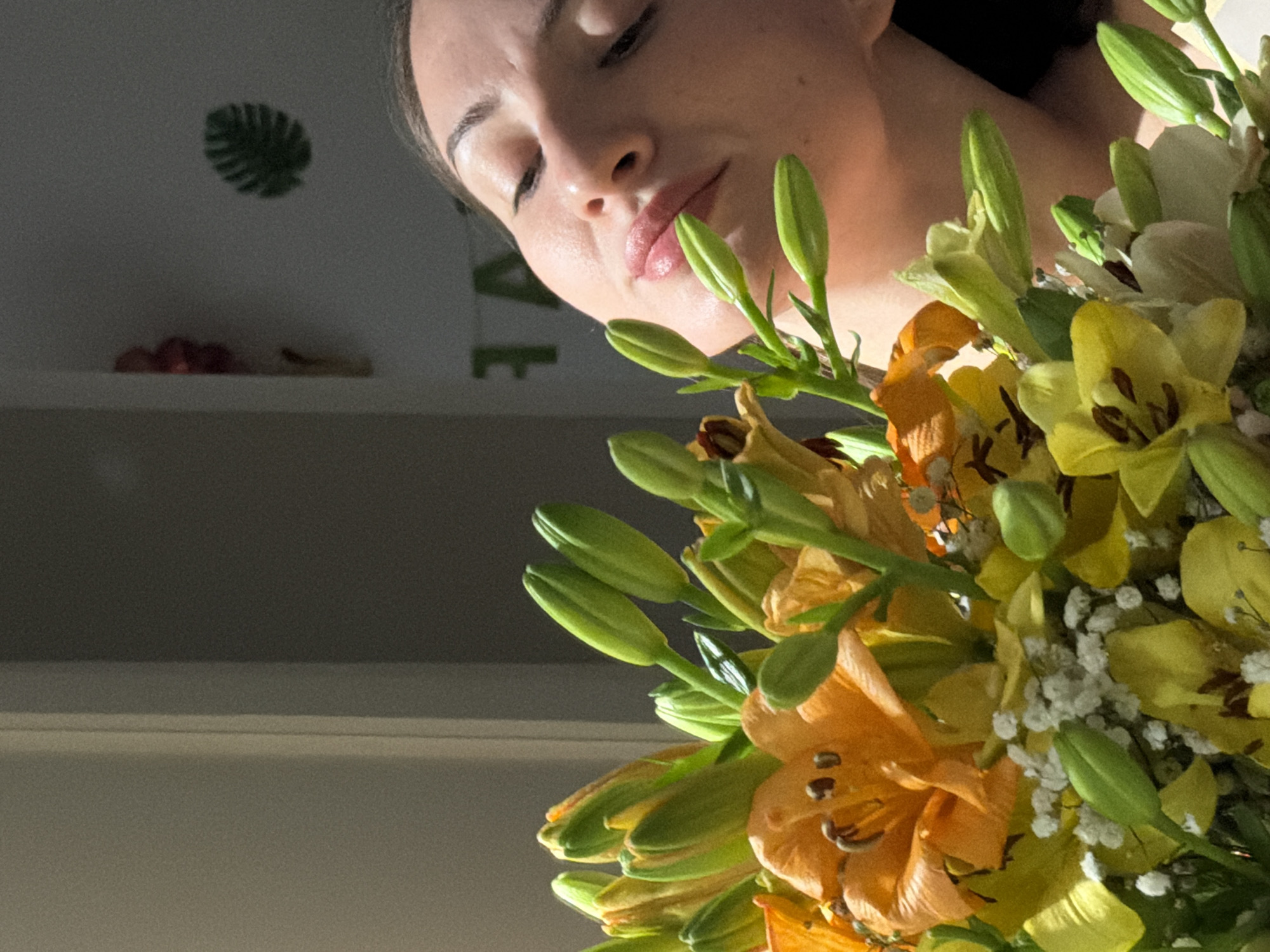 The image shows a close-up of a bouquet of lilies, featuring various shades of orange, yellow, and white, interspersed with green buds and stems. In the background, a woman's face is partially visible, with her eyes closed and her head tilted back, seemingly enjoying the scent of the flowers. The lighting suggests an indoor setting, possibly a room in Sharm El Sheikh, Egypt, with natural light entering from the left. Decorative elements, such as a leafy wall decal and a cluster of red balloons, are visible on the wall behind the woman, hinting at a festive or decorative ambiance. The scene captures a moment of serene enjoyment, with the vibrant colors of the flowers contrasting with the soft focus on the woman and the muted background.