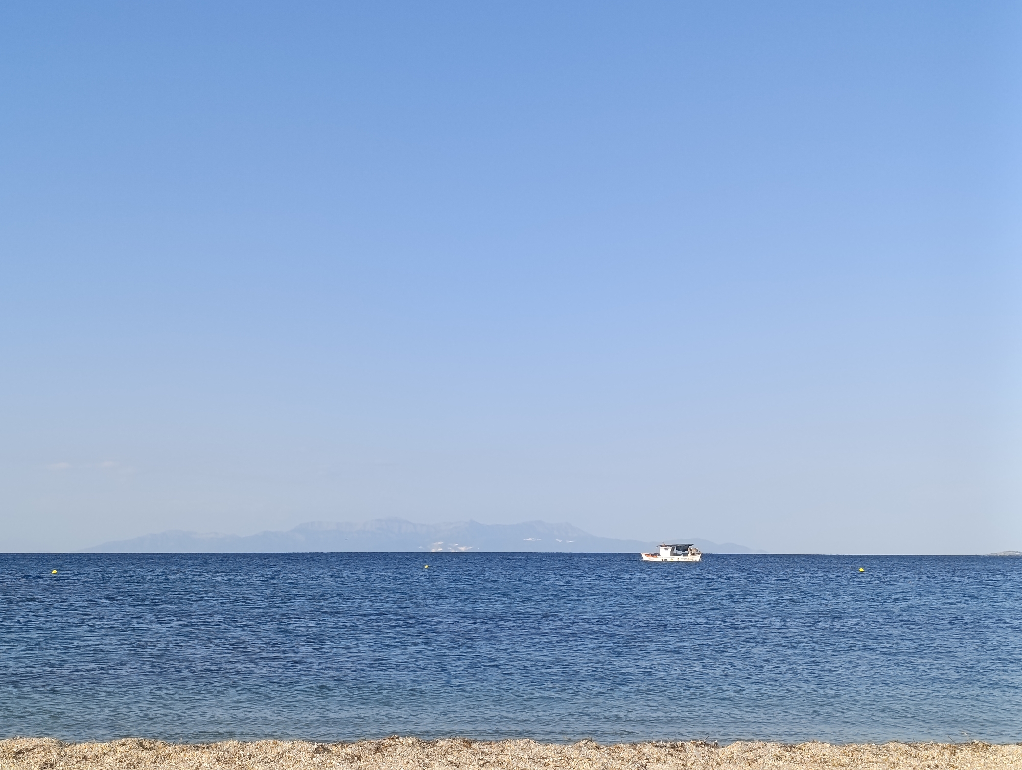 The image depicts a serene seascape with a clear blue sky and calm, deep blue water. In the distance, a range of hazy mountains forms the horizon line. A small white boat with a dark roof is visible on the right side of the water. The foreground features a pebbly beach. The scene appears to be located in the city of Fanari, Greece, suggesting a coastal view. There are no people visible in the image. The environment is tranquil, with minimal activity shown. The lighting suggests it is daytime, with the sun high in the sky.