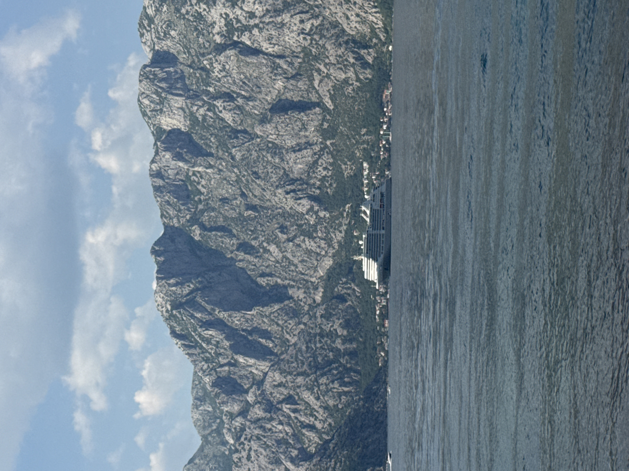 The image depicts a cruise ship docked near the town of Prčanj, Montenegro, situated along the Bay of Kotor. The ship is positioned close to the shore, with a backdrop of steep, rocky mountains covered in vegetation and partially obscured by low clouds. The water is calm, with visible ripples and reflections. The sky is blue with scattered white clouds. Buildings and other urban structures are faintly visible on the shoreline between the ship and the mountain.