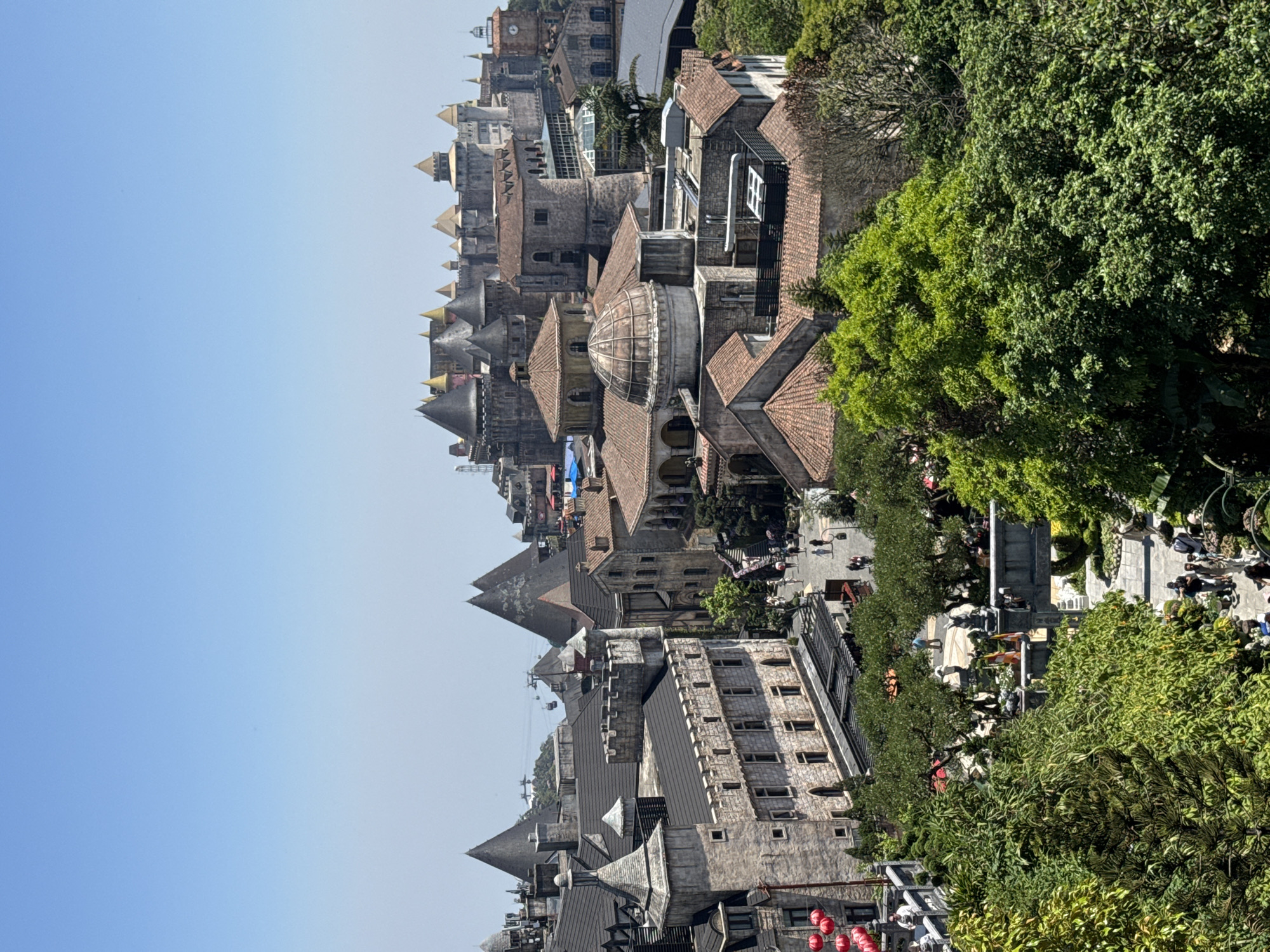 The image captures a high-angle view of the European-style architecture at Ba Na Hills in Đà Nẵng, Vietnam. Several buildings with red-tiled roofs, domes, and pointed towers are visible. A clear, blue sky is on the left. There are trees on the right of the image. In the middle of the image, a populated walkway area shows people moving around.