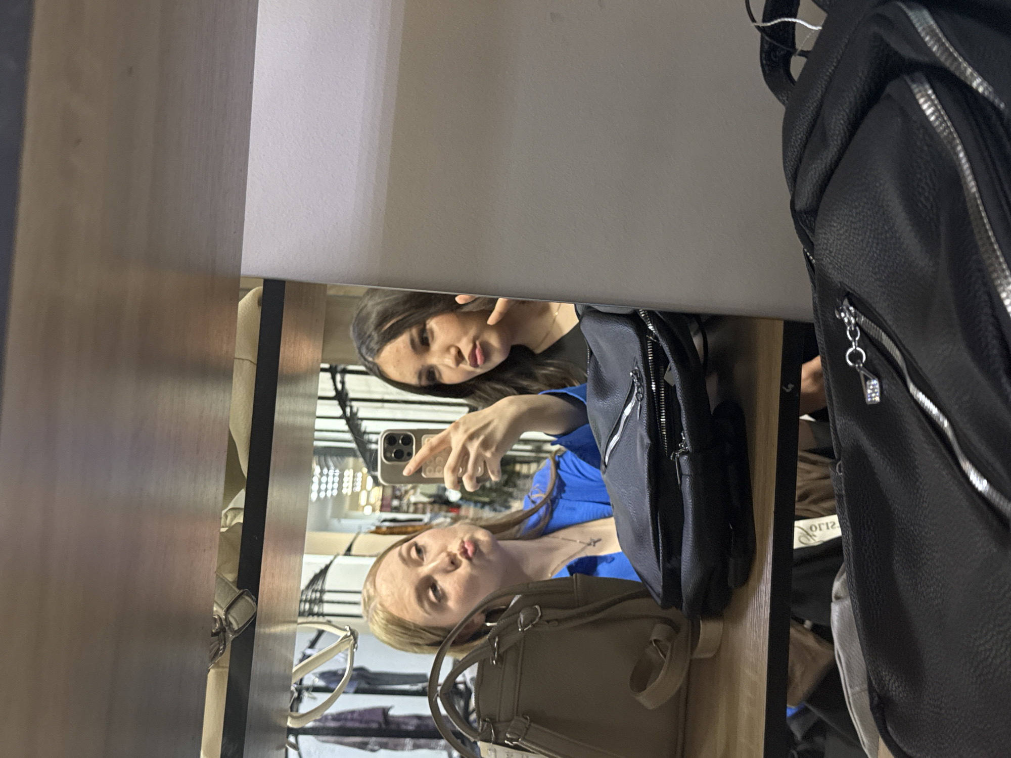 The image, taken in a retail environment resembling a clothing store, captures a mirror reflection of two young women. The woman on the left, with blonde hair, is wearing a bright blue shirt and a cross necklace. She is holding a smartphone and taking a selfie, puckering her lips. Next to her, a woman with dark brown hair, wearing a dark top, is also looking at the phone, with her lips pursed and a finger near her mouth. In the foreground, several bags are visible, including a taupe handbag and a black backpack with silver zippers. The background, reflected in the mirror, shows clothing racks and bright ceiling lights, suggesting a shopping mall or department store setting. The overall impression is a candid moment of friends taking a picture while shopping. There is no specific indication of the city of Perm, Russia.