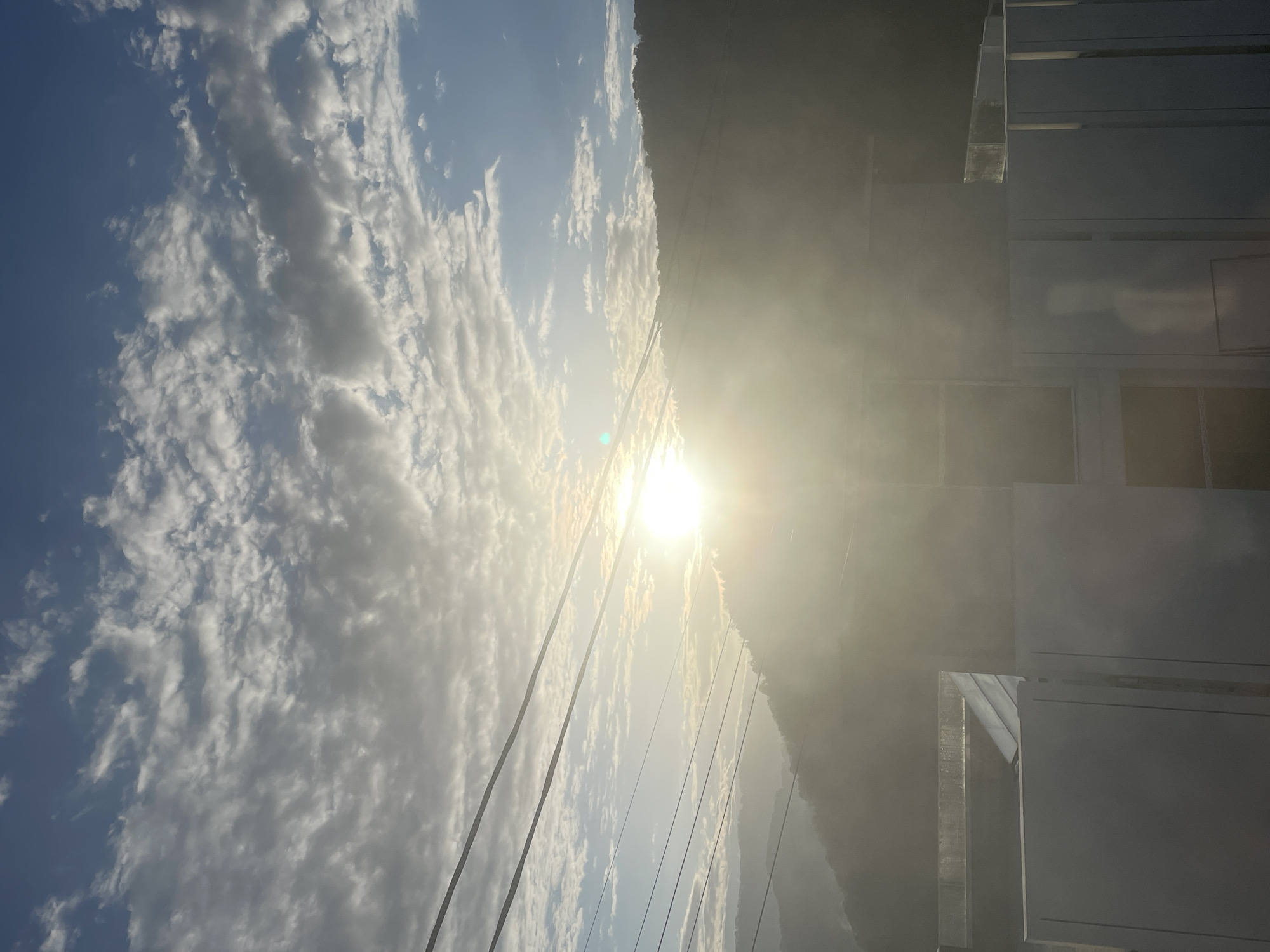 The image shows a bright, sunlit sky with scattered cumulus clouds above a dark mountain range. Overhead power lines cut across the view. To the right of the frame is a white, geometric structure of a building or structure. The image is rotated on its side. The location in the city of Baranovka, Russia is unknown.