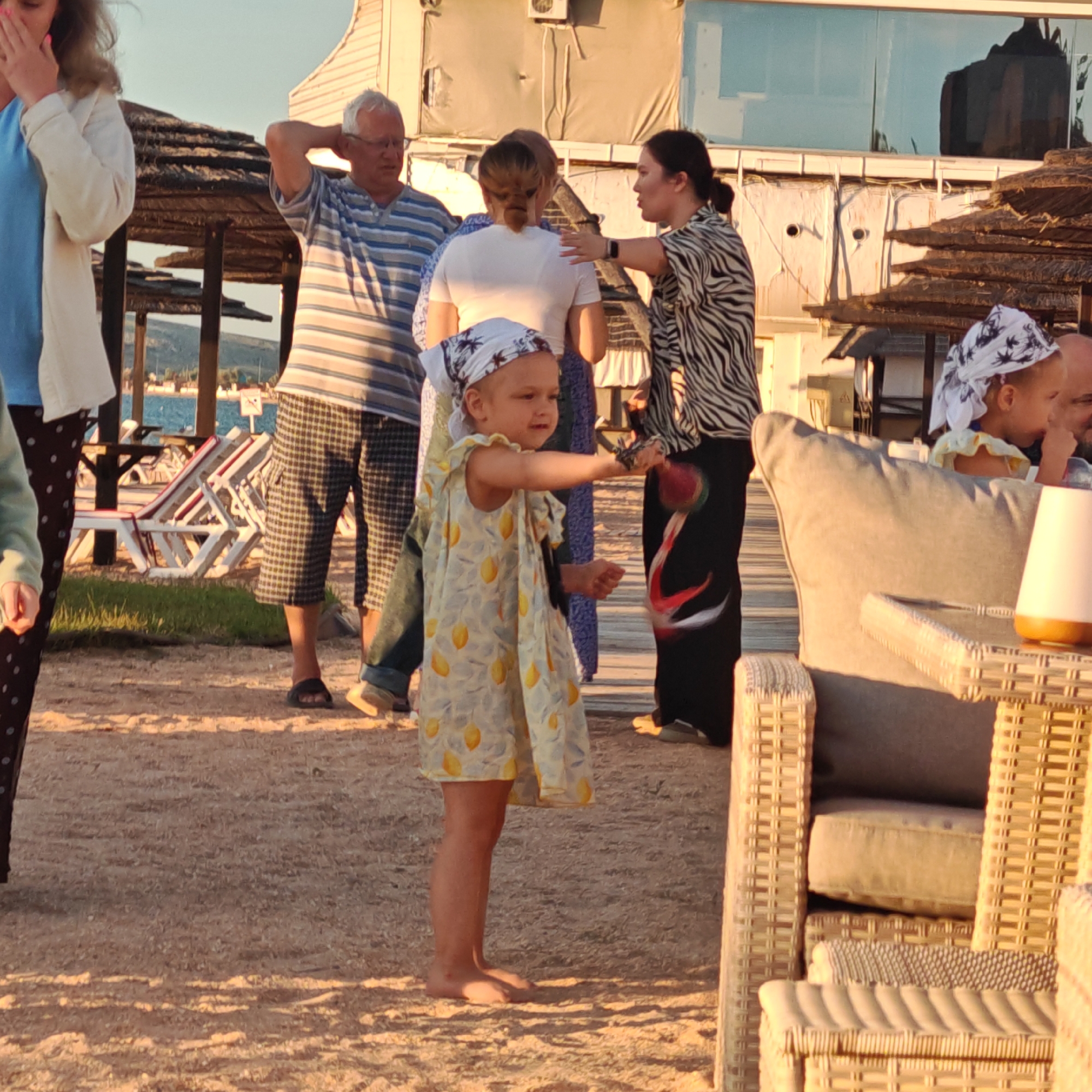 In the city of Shchelkino, Ukraine, a group of people are gathered on a sandy area, possibly a beach or a promenade. In the foreground, a young girl with a bandana on her head and a yellow dress adorned with lemons is actively playing, holding a red and black toy. Behind her, an adult woman in a black and white zebra-print top and black pants is looking towards the girl. To the left, a man with glasses and a striped shirt is visible, and further back, a woman in a white shirt. The environment includes thatched sunshades, lounge chairs, and a building with large windows in the background. The lighting suggests it is late afternoon or early evening, with warm sunlight casting shadows.