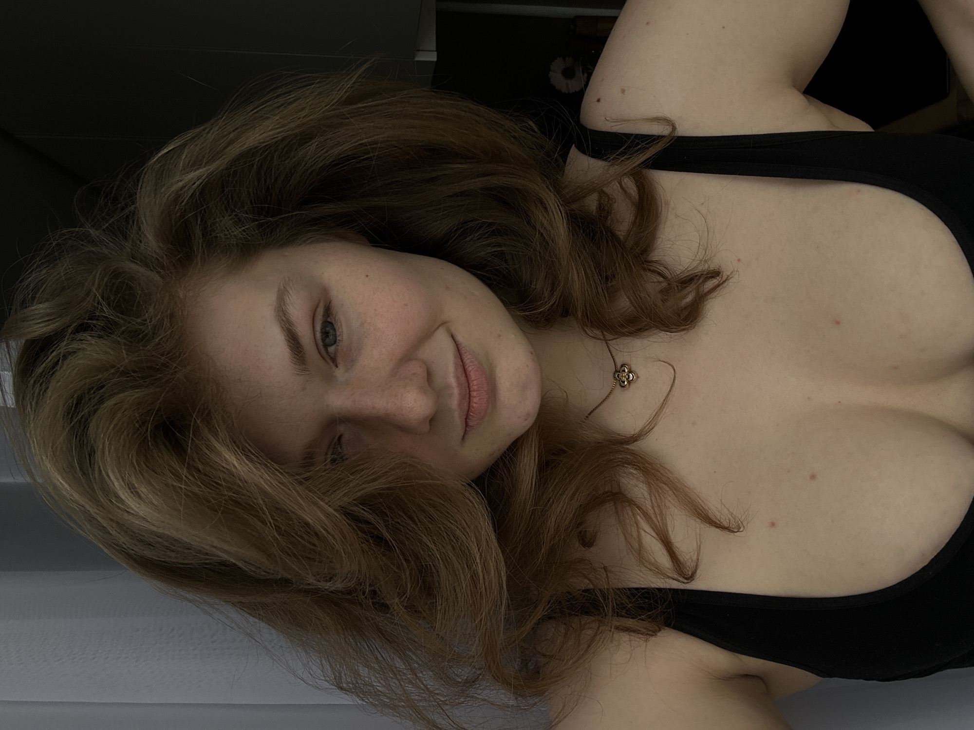 A woman with light brown hair and freckles is lying down wearing a black tank top and a gold necklace with a floral pendant. She is angled to the left and smiling slightly. The background is dark and out of focus. The location in the city of Kazan, Russia, cannot be determined from the provided image.