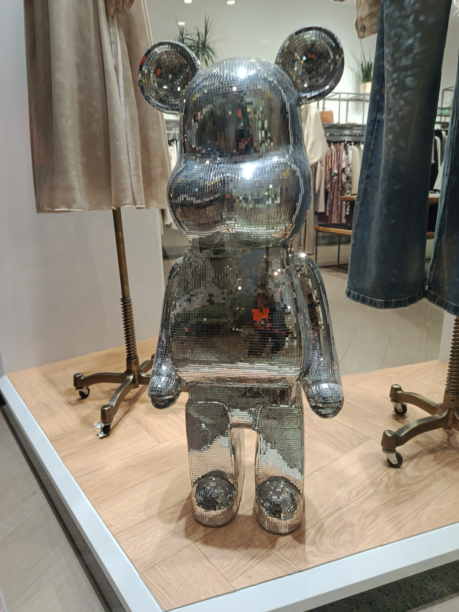 The photograph depicts a silver, mirrored teddy bear figure in a retail environment. The bear is positioned on a light wood-grain platform with a white border, suggesting a display or staging area. Clothes on hangers are visible in the background, implying a clothing store setting. The lighting is bright and diffuse. The specific location in Moscow, Russia, cannot be determined from the image alone.