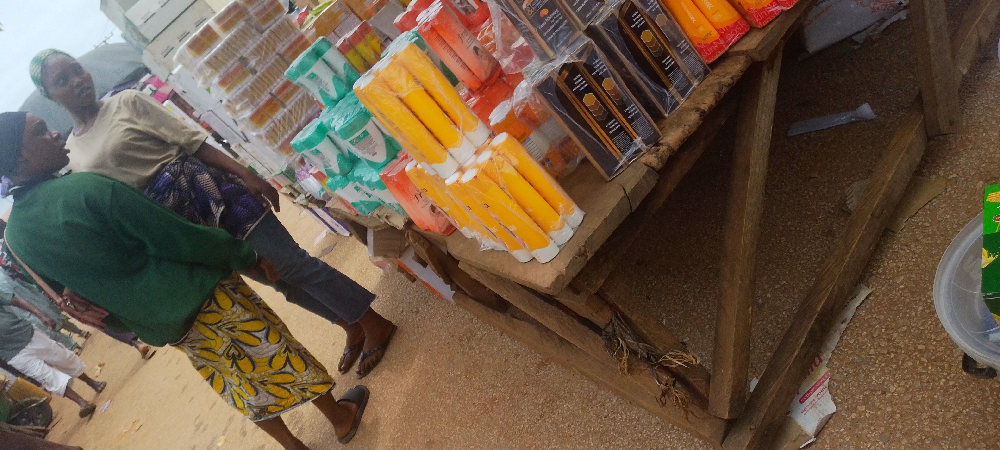 In Baissa, Nigeria, a market scene unfolds with a close-up view of two women interacting near a wooden stall laden with various consumer goods. The woman on the left, wearing a green long-sleeved shirt and a yellow and white patterned skirt, is positioned in the foreground. Her dark hair is covered with a headscarf. To her right stands another woman, wearing a light brown t-shirt and a patterned blue and white skirt, her head also covered with a headscarf. They appear to be engaged in a conversation, with their bodies oriented towards each other.
The stall, constructed from rough wooden planks, displays an assortment of products, predominantly cosmetics and toiletries, neatly packaged and stacked. Prominent among these are rows of yellow cylindrical tubes, some wrapped in plastic, and orange boxes with visible branding such as "Frya." Other items include green packaged products and dark boxes with text indicating "Intensive Toning" and "Solaray." The ground surface is composed of gravel and sand, typical of an outdoor market environment. The overall lighting suggests an overcast day. The image captures a candid moment within a bustling commercial setting, highlighting everyday life in Baissa.