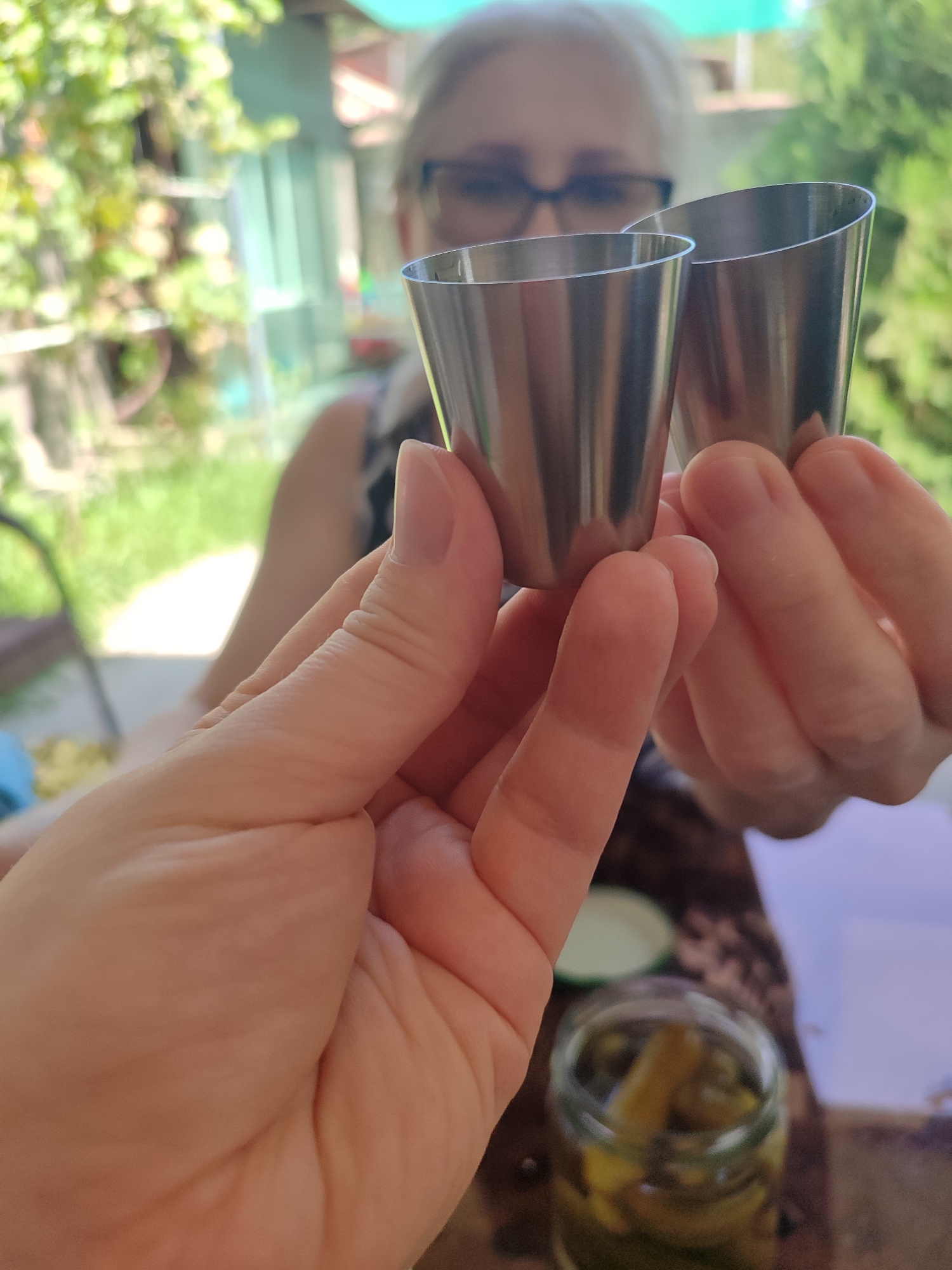 In Eshmataul, Uzbekistan, two hands are holding up two small metal shot glasses in a toast. The person in the background, slightly out of focus, is a woman with blonde hair wearing glasses and a patterned top. She is likely holding one of the shot glasses, out of frame. The foreground shows the hands holding the glasses. In the bottom right, a glass jar containing pickles is visible on a table. The background is blurred, showing greenery and structures suggestive of an outdoor or garden setting.