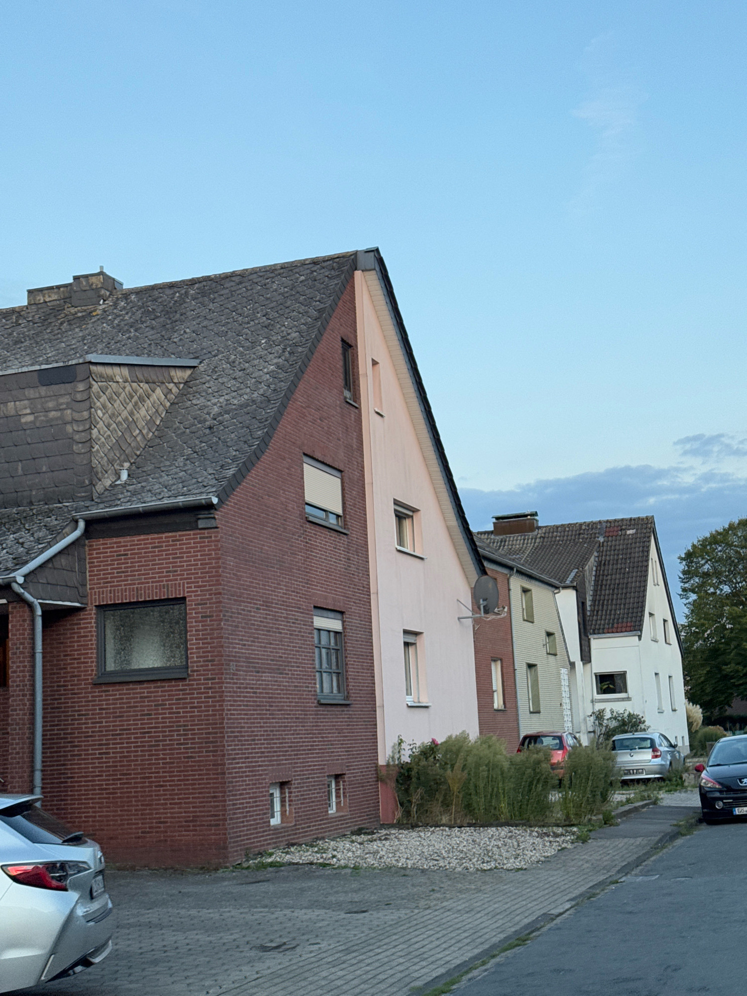 The image shows a row of houses on a street in Cleves, Germany. The houses are of varying styles and colors, with pitched roofs and visible windows. Several cars are parked on the street and in driveways. The foreground shows a silver car and a paved driveway. In the background, a large tree is visible. The sky is clear with a few clouds. The scene appears to be during the daytime.