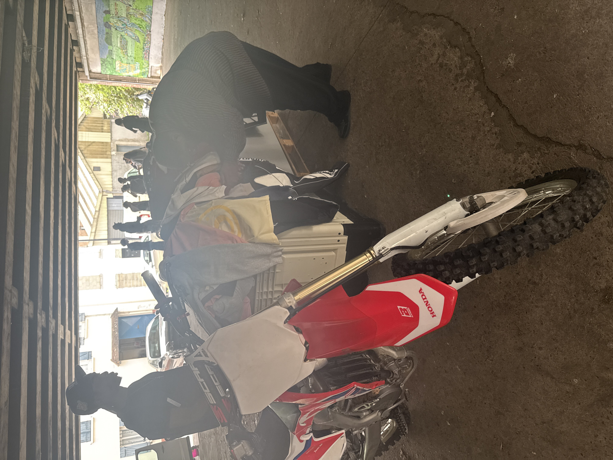 Underneath a concrete overpass in Saint-Denis, France, a person wearing a black baseball cap and a black t-shirt stands next to a white and red dirt bike. The dirt bike is facing left, with its front wheel towards the camera. Behind the dirt bike, a white pallet is piled high with clothing. To the right of the pallet, another person, wearing a dark striped sweater and black pants, is bent over, appearing to sort through the clothing. In the background, several other people are visible, and a mural depicting a landscape scene is painted on a wall to the far right. Buildings and trees can be seen beyond the overpass.