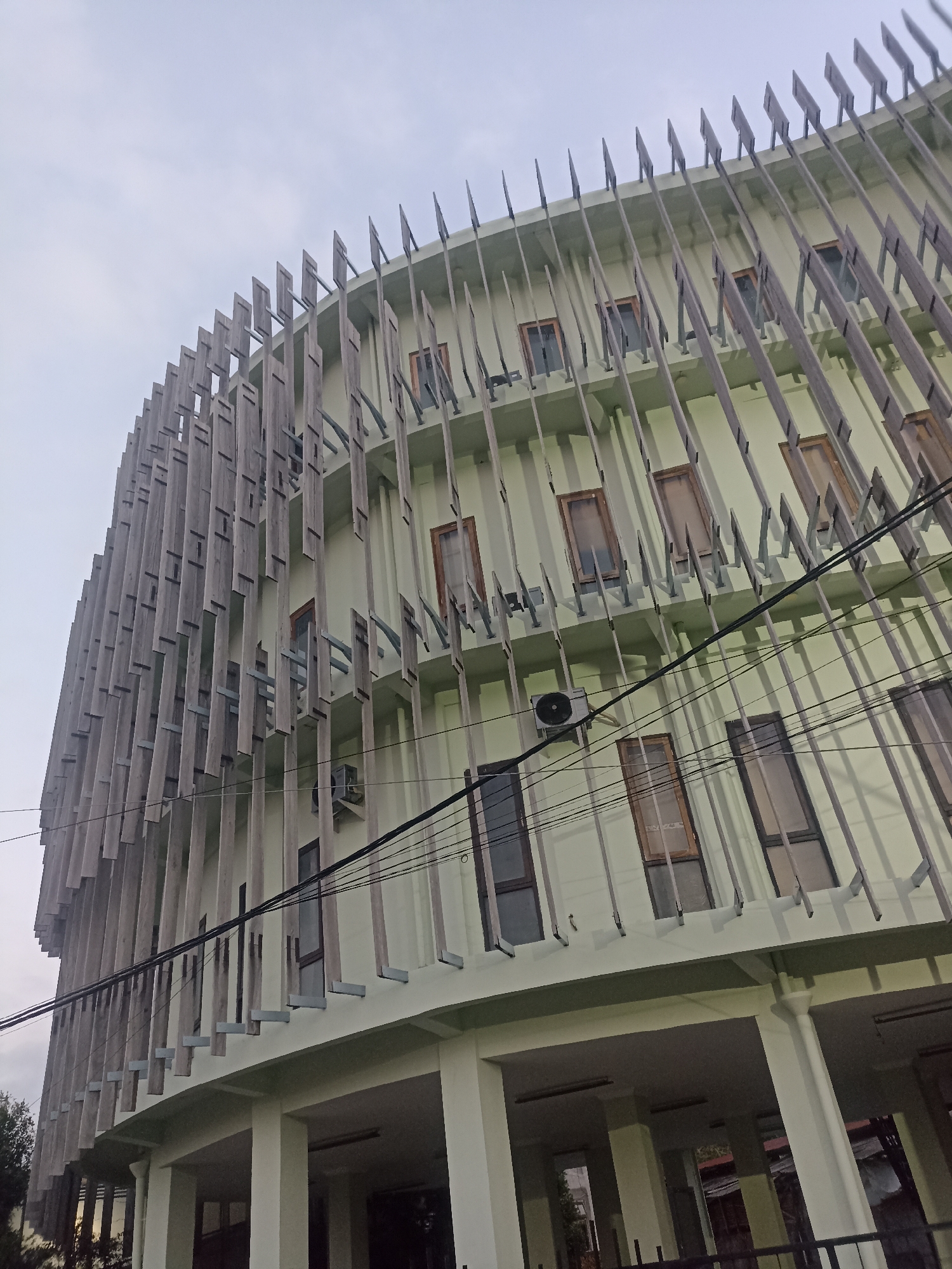 This is a low-angle outdoor photograph of a light green, multi-story building with a unique facade. The building's exterior features vertical wooden slats, angled and spaced out, creating a textured and rhythmic pattern across multiple floors. Behind these slats, rectangular windows with dark frames are visible, some with a wooden trim. A portion of an air conditioning unit is mounted on the building's wall, and electrical wires crisscross diagonally across the facade. The lower level of the building has a covered walkway supported by white columns. The sky above is overcast and pale. The scene appears to be located in Mojopanggung, Indonesia.
