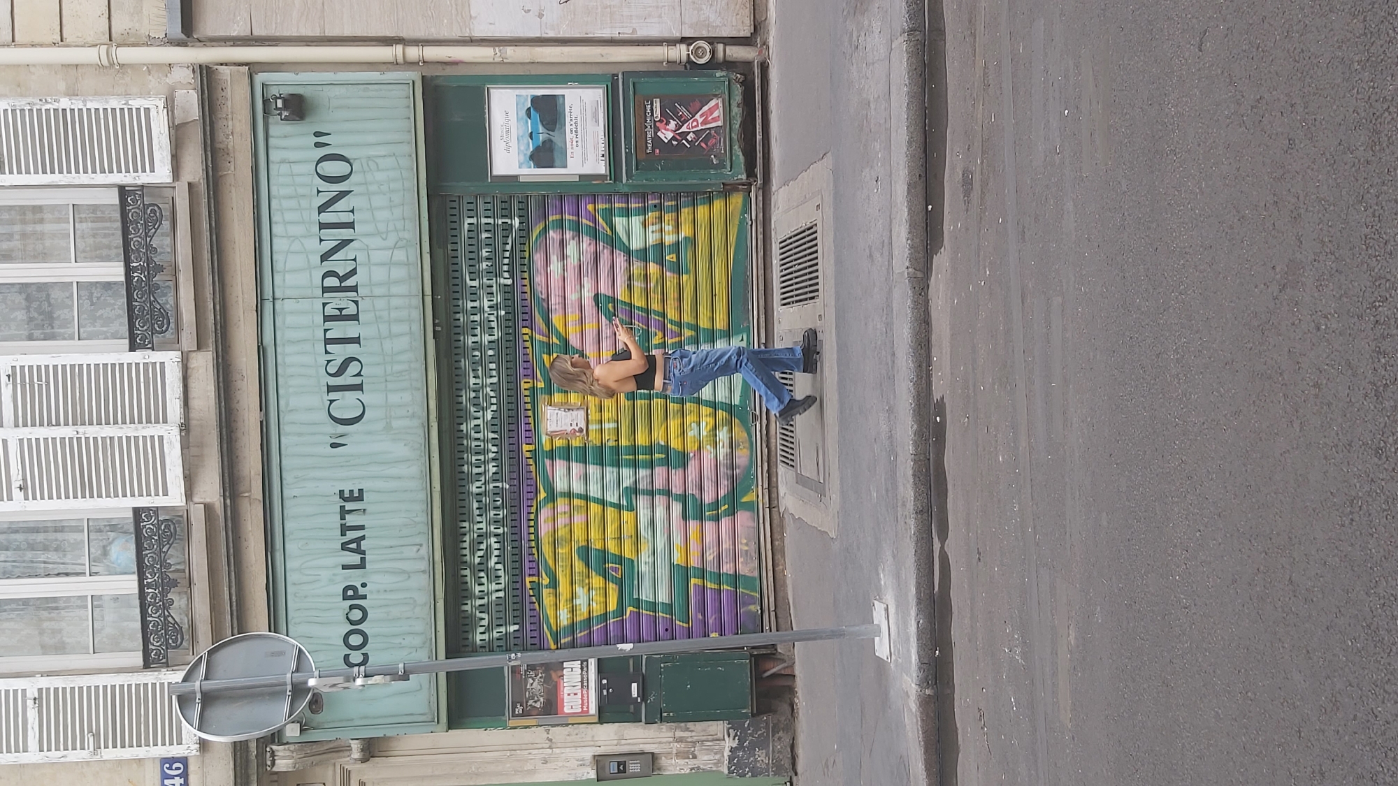 A woman is walking on a sidewalk in front of a closed store in Paris, France. The store has a teal-colored facade with the words "COOP. LATTE" and "CISTERNINO" written on it. The store's rolling metal security gate is covered in colorful graffiti. The woman is wearing a black crop top, blue jeans, and black boots. She is looking at her phone as she walks. There are two posters on the wall next to the store entrance. One poster has the word "diplomatique" at the top. The other poster advertises "THEATRE MICHEL". The sidewalk is paved with stone and there is a street in front of the sidewalk. The building above the store has windows with white shutters. The number "46" is visible on the building to the left.