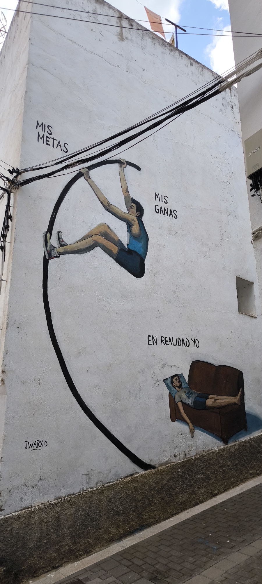 The image displays a mural painted on the exterior wall of a building in Fanzara, Spain. The mural depicts a pole vaulting athlete suspended in mid-air, with the text "MIS METAS" (My Goals) above and "MIS GANAS" (My Desires) to the right of the athlete. Below the pole vaulter, the text "EN REALIDAD YO" (In reality, me) is written, accompanied by a painting of a man lying on a brown couch. The artwork is signed "J.WARX :)" in the lower left corner. The building wall is white and textured, with visible cracks. Black electrical wires run across the top of the mural. In the background, a blue sky with scattered clouds and some orange fabric flags are visible. The lower portion of the image shows a paved street and a textured stone wall.