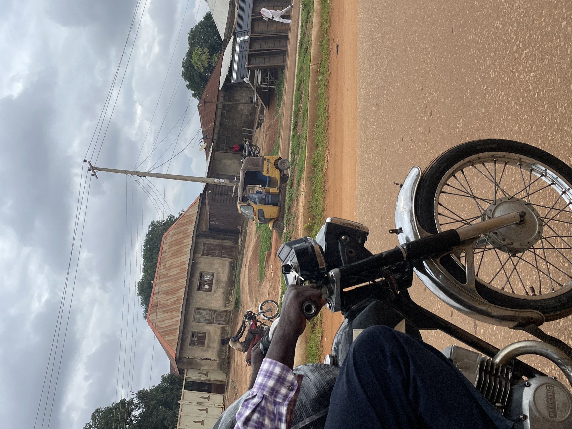 The image captures a street scene in Minna, Nigeria, under a cloudy sky. In the foreground, a person's hands, wearing a plaid shirt, grip the handlebars of a motorcycle. The motorcycle's front wheel and part of its engine are visible. In the middle ground, several buildings with corrugated iron roofs line the street. A yellow three-wheeled auto-rickshaw is parked on the side of the road. A person on a red motorcycle rides past in the distance, and another person in white attire walks further down the street. A wooden utility pole with electrical wires stands prominently on the right side of the frame. The ground is a reddish-brown dirt road.