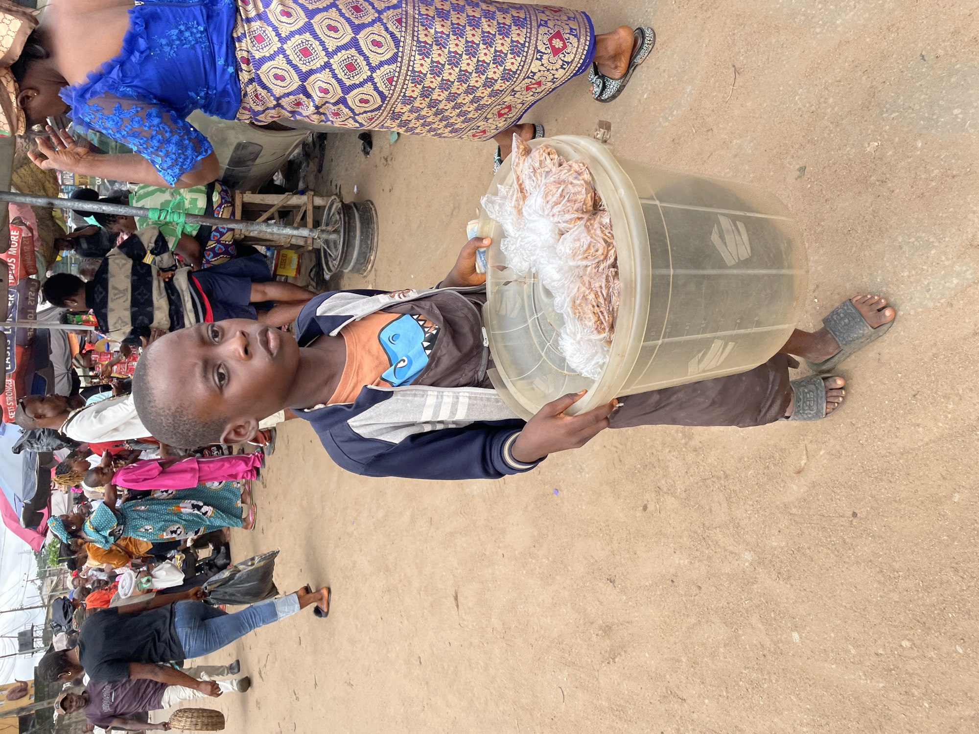 In an outdoor marketplace scene in an unknown city, a young boy is the central subject, holding a large clear plastic tub filled with bagged snacks. He wears a dark blue and grey jacket over an orange shirt with a blue dinosaur graphic. The boy's dark brown skin and focused expression are prominent. He stands on a dirt ground, wearing dark grey slides. To his right, a woman, partially visible, wears a bright blue, sheer top with intricate lace details and a vibrant, patterned skirt. Her feet are adorned with patterned black and white slides. In the background, a bustling market environment is suggested with various people and stalls. Several individuals are seen walking and standing, some carrying bags and baskets. Overhead, market umbrellas of red and pink hues provide shade. The ground is a mixture of dirt and sand, with scattered debris. The overall atmosphere is one of lively commerce.