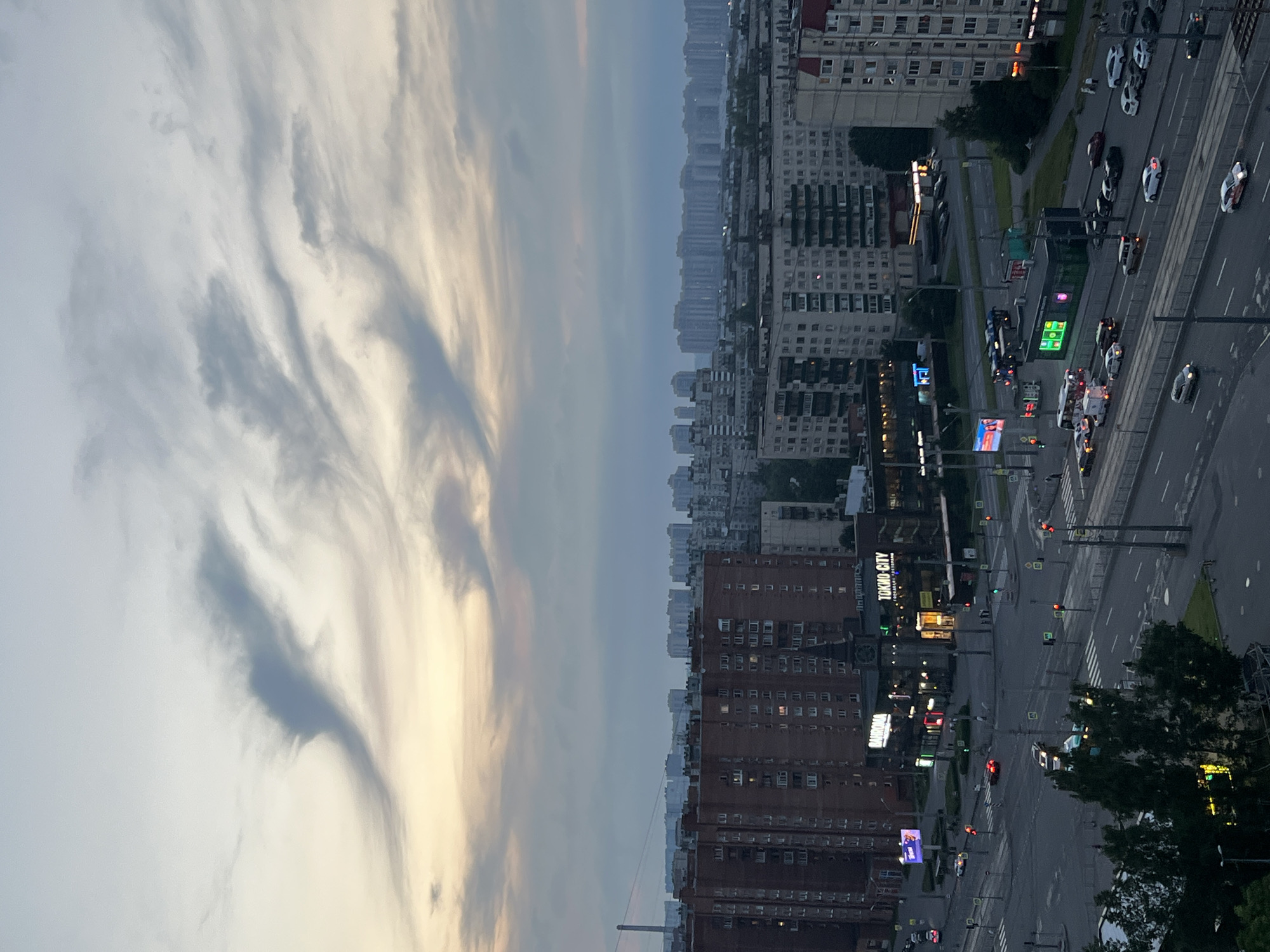 The image captures an aerial view of a city intersection at dusk, likely in an urban area of Russia, given the Cyrillic signage. The sky is a mix of soft yellow and gray clouds, with wispy formations high up. Below, a dense cityscape stretches into the distance, characterized by numerous apartment buildings of varying heights and architectural styles, predominantly in muted tones of brick, concrete, and light gray.
In the foreground, a multi-lane road curves through the scene, with cars and buses in motion. Traffic lights are visible, indicating active movement at the intersection. Several commercial buildings with illuminated signage are situated along the road. One prominent sign reads "TOKIO CITY," and another appears to be for "BARKHOMA." There are also smaller kiosks and streetlights contributing to the illuminated urban landscape. Pedestrians can be seen on the sidewalks. The overall impression is one of a functional, albeit somewhat monotonous, urban environment transitioning into evening.