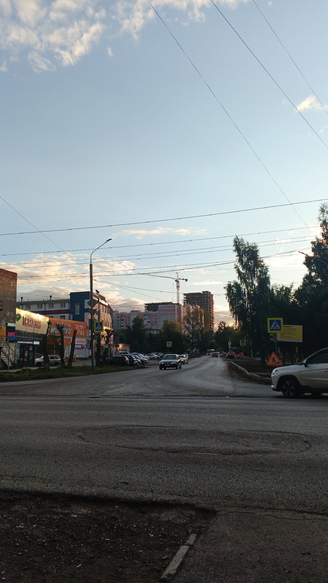 The image captures a street scene in Izhevsk, Russia, during what appears to be late afternoon or early evening, indicated by the warm lighting and long shadows. The perspective is from a roadside, looking down a multi-lane road that curves to the right.
In the foreground, the road surface is visibly worn with patches and cracks. To the left, a row of buildings lines the street. These buildings include shops with bright signage, some with their names partially obscured but suggesting commercial activity such as "Magnit" and a general store. A street lamp stands prominently on the sidewalk.
Further down the road, construction is evident with several multi-story buildings under various stages of completion, including one with a prominent crane. Trees line the right side of the road, casting shadows onto the pavement.
Several vehicles are present: a white car is in the right lane, while other cars are on the road and parked along the left side. Pedestrian crossing signs are visible, indicating an intersection. The sky above is a pale blue with scattered white clouds, some tinged with warm light. Overhead power lines crisscross the frame. The overall impression is of a moderately busy urban environment with ongoing development.