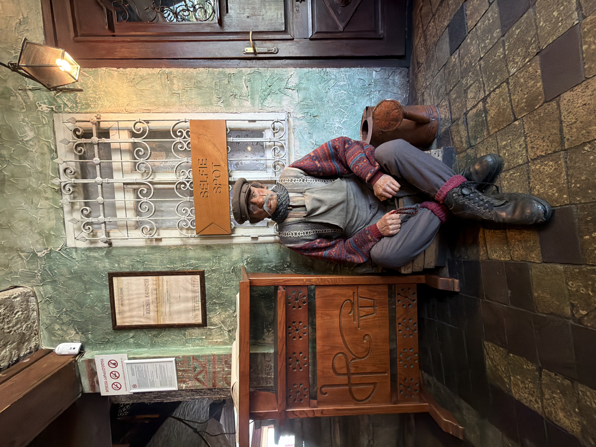 This image depicts a man, likely a mannequin, seated on the floor in front of a window with decorative bars. He is wearing a cap, a scarf, overalls, a sweater, and boots. A string of beads is held in his right hand. To his left is a wooden cabinet with carved designs. Above the mannequin, a wooden sign reads "SELFIE SPOT." The background features a textured green wall, a window, and a wooden door. The floor is tiled with dark, irregularly shaped stones. This scene is located in Tbilisi, Georgia, and appears to be inside a building, possibly a restaurant or shop, given the signage and the display of what looks like a framed certificate on the wall.