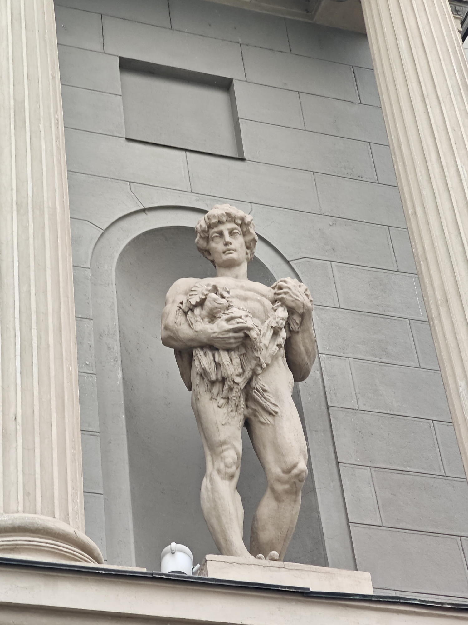 A low-angle, full-shot photograph captures a stone statue of a muscular man against a grey, stone-paneled building. The statue is positioned in a niche and is flanked by two large, fluted columns. The man in the statue has curly hair and is holding a bundle of foliage or branches in his arms, which are crossed in front of his chest. He is depicted with a muscular physique, wearing what appears to be a loincloth made of leaves. His stance is dynamic, with one leg slightly forward, suggesting movement.
The building behind the statue is constructed from large, rectangular grey stone blocks, with a subtle recessed rectangular panel above the niche. The architectural style suggests a classical influence, with the imposing columns and the overall grandeur of the structure. A white, modern-looking cylindrical object, possibly a light fixture or camera, is visible on the ledge beneath the statue.
The lighting appears diffused, indicating an overcast day or possibly late afternoon. There are no clear indicators of a specific time of day or weather conditions beyond the soft light. The image provides a sense of scale and solidity, with the ancient-looking statue contrasted against the backdrop of the building. The location context provided is Moscow, Russia, though no specific landmarks are identifiable within the frame. The overall mood is one of solemnity and classical artistry.