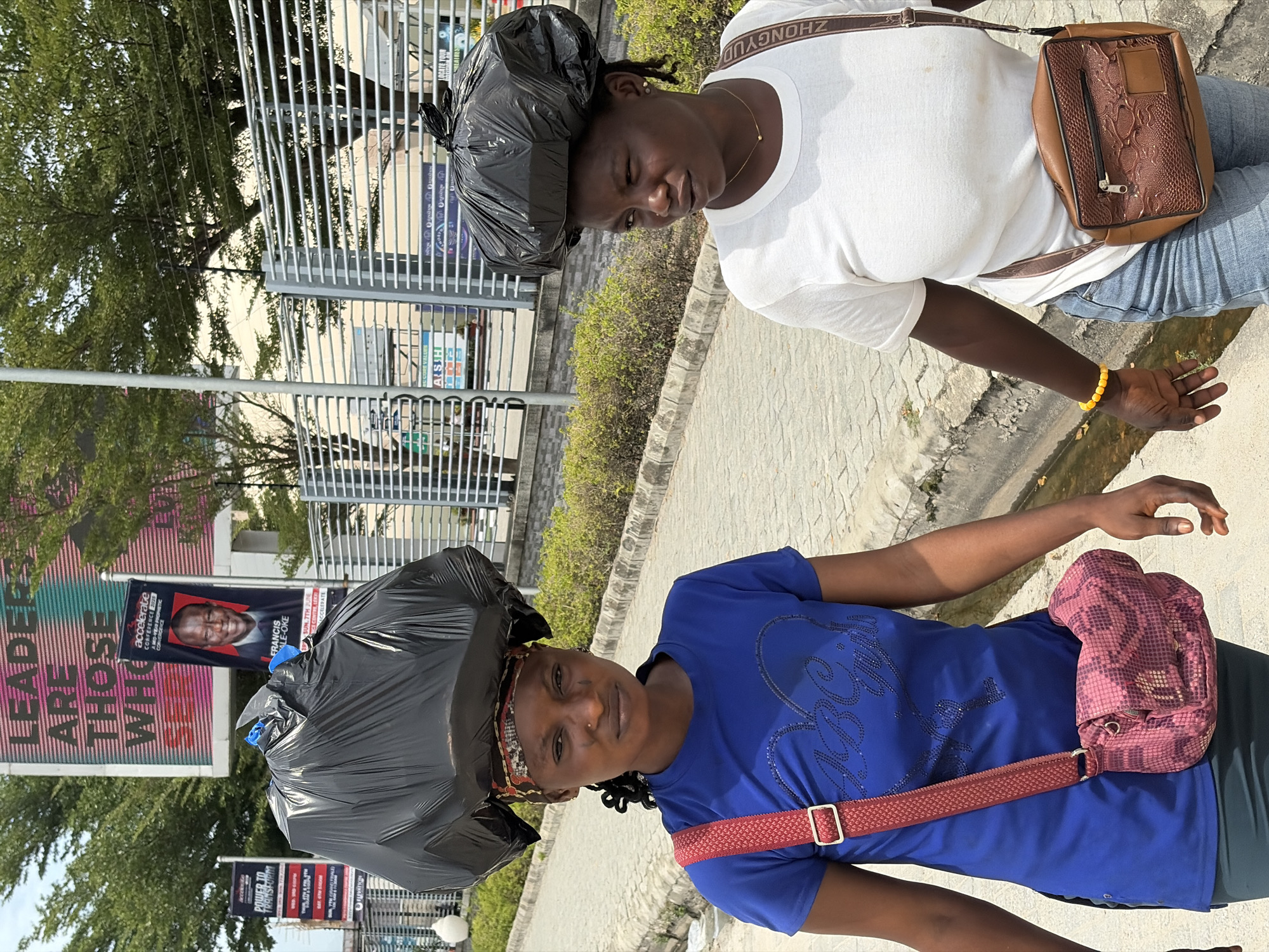 Two women stand on a sidewalk in Igboefon, Nigeria. Each has a large black plastic bag on her head, seemingly as protection from the elements. The woman in the foreground wears a blue shirt and a patterned bag across her body. Behind her, a woman in a white shirt wears a brown bag with a snakeskin pattern. In the background, there are buildings with metal bars and campaign posters, including one for a "Francis Ale-Oke" conference and another stating "Leaders are those who...". The environment includes concrete sidewalks, green foliage, and utility poles.