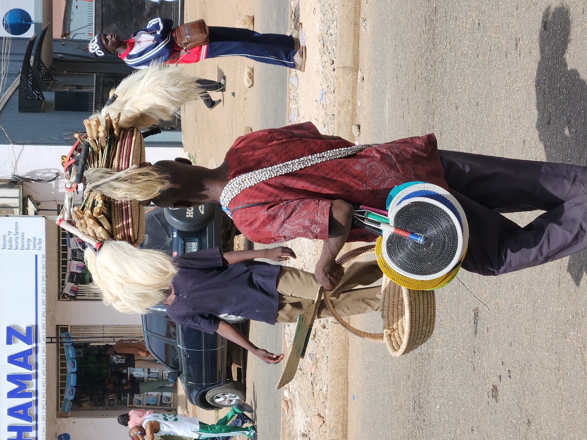 The image shows a city street in Unknown, Unknown, where a man is selling handmade woven fans and baskets. He wears a red shirt with a string of white beads around his neck and dark pants. The man carries a stack of colorful woven fans and has a woven basket hanging from his arm.
Behind him, a younger person with blonde hair, dressed in a blue shirt and khaki pants, walks with a similar woven basket. In the background, there is a store with the name "HAMAZ" prominently displayed. Several people can be seen in front of the store. Another man with a white headdress and a fuzzy white accessory walks on the opposite side of the street, wearing a blue and red outfit with a brown bag. A black car is parked on the side of the road. The street appears to be paved and is exposed to bright sunlight, casting shadows of the figures in the image.