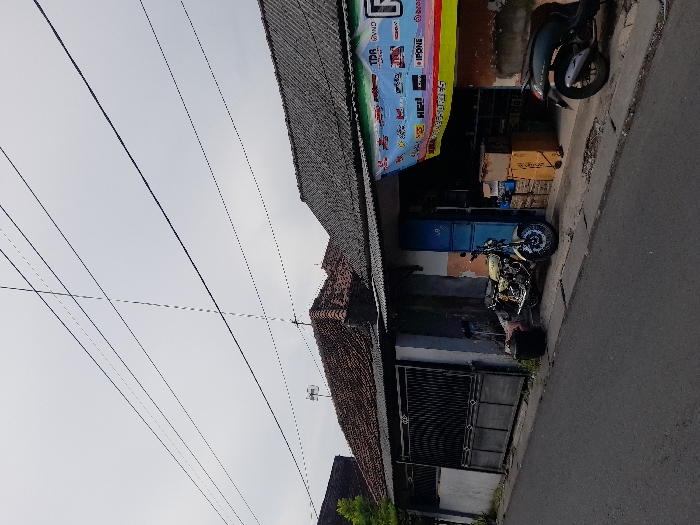 The image shows a street in Kecamatan Tanggulangin, Indonesia (latitude: -7.505, longitude: 112.716). The street is paved and runs from left to right in the image. There is a house on the right side of the street. The house has a tiled roof and a concrete facade. There is a large, black metal gate in front of the house. The house has a shop front with a blue door and windows. There is a sign above the shop front with the words "TDR" and "WIND" on it. A yellow motorcycle is parked in front of the shop. A black scooter is parked in front of the shop on the right side of the image. There are several black power lines strung overhead across the image. The sky is overcast. There is no visible vegetation in the immediate foreground.
