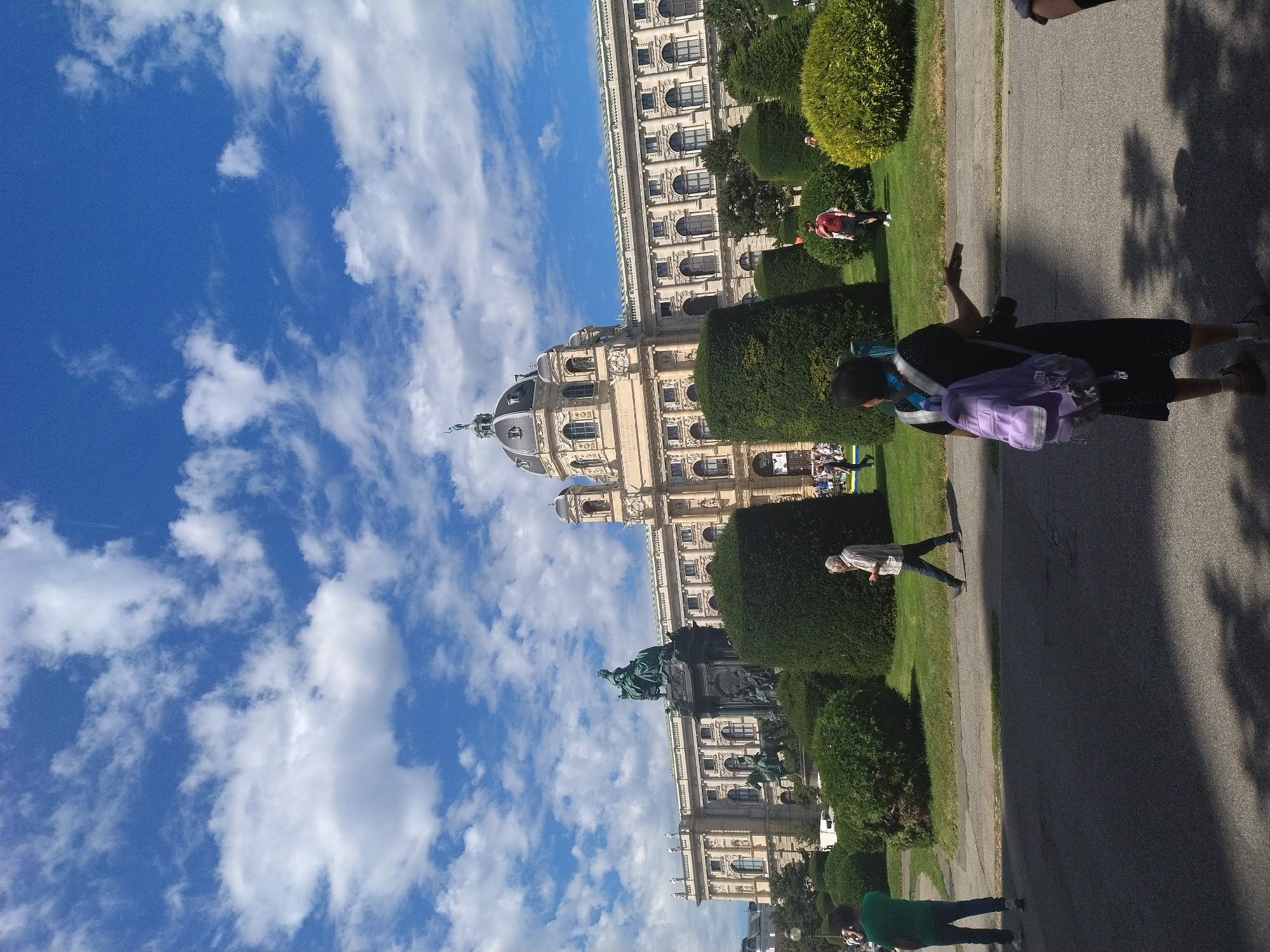 The image depicts the exterior of the Naturhistorisches Museum in Vienna, Austria, on a sunny day with a blue sky and scattered clouds. In the foreground, a woman with a purple backpack and a camera is visible from the back, facing away from the viewer. To her left, a person in a green shirt and jeans is standing. To the right of the woman, a man in a checkered shirt and dark pants is walking away from the viewer.
Behind the people, there is a meticulously manicured garden with large, spherical hedges. Further in the background, the grand facade of the Naturhistorisches Museum is visible, featuring classical architecture, numerous arched windows, and a prominent dome. A large bronze equestrian statue is situated in front of the museum. Several other individuals can be seen walking in the distance near the museum entrance. The overall scene is a public space, likely a park or square in front of a significant cultural institution.