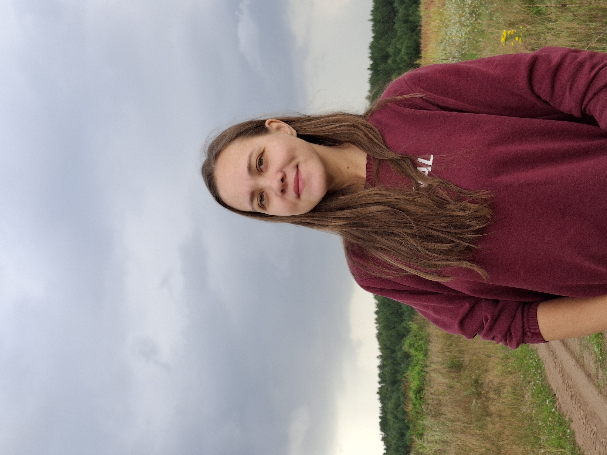 A medium shot shows a young woman with long, brown hair standing outdoors. She is wearing a maroon sweatshirt with white lettering that reads "VAL" on the chest. The woman is smiling slightly and looking at the camera. She is positioned in front of a field of tall, dry grass and wildflowers, with a dirt path visible to her left. A line of dark green trees forms the horizon behind her, and the sky above is overcast with gray clouds. The overall setting appears to be a rural area, potentially the city of Plishkino, Russia, given the information provided.